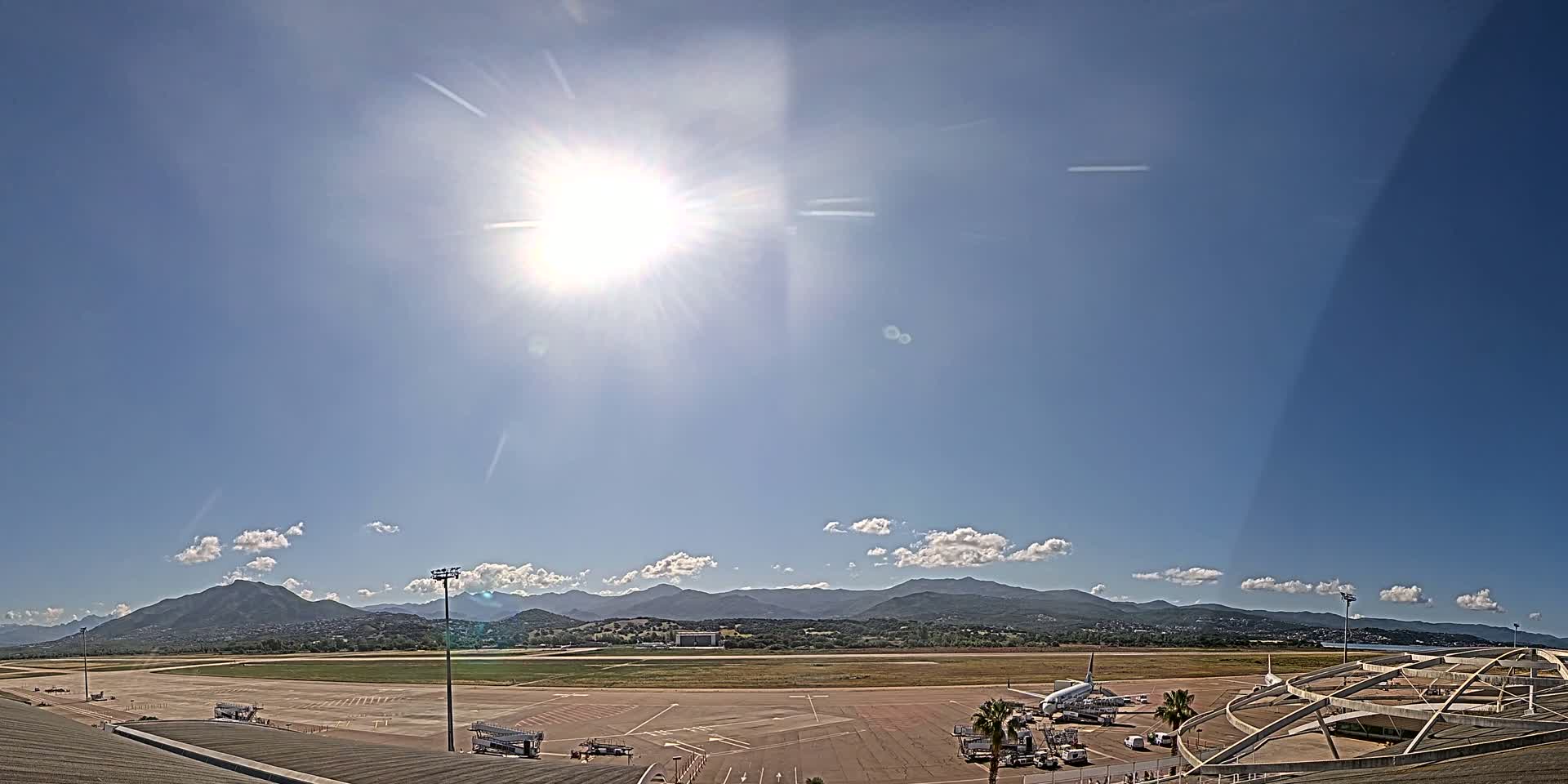 A sunny day at an airport shows a passenger plane parked at a gate with mountains in the background.