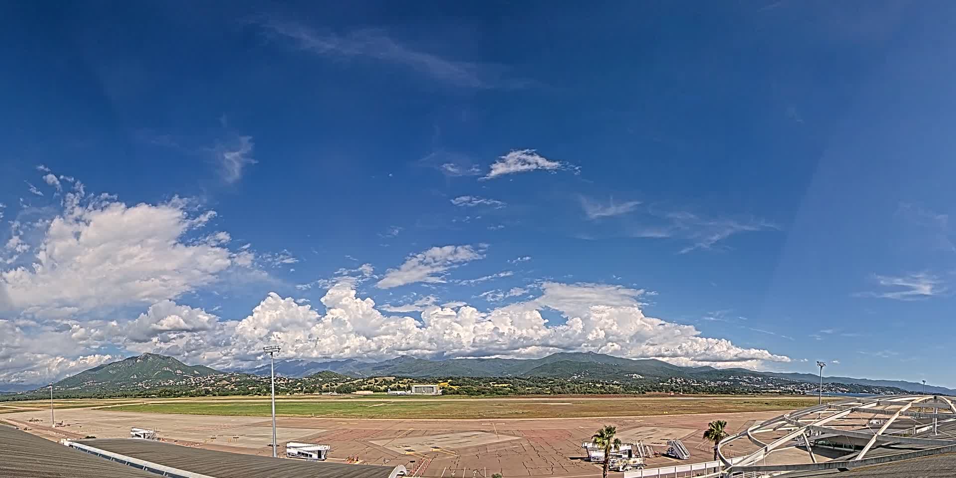 A mostly sunny day with puffy cumulus clouds over a large airport tarmac and runway, backed by distant mountains.
