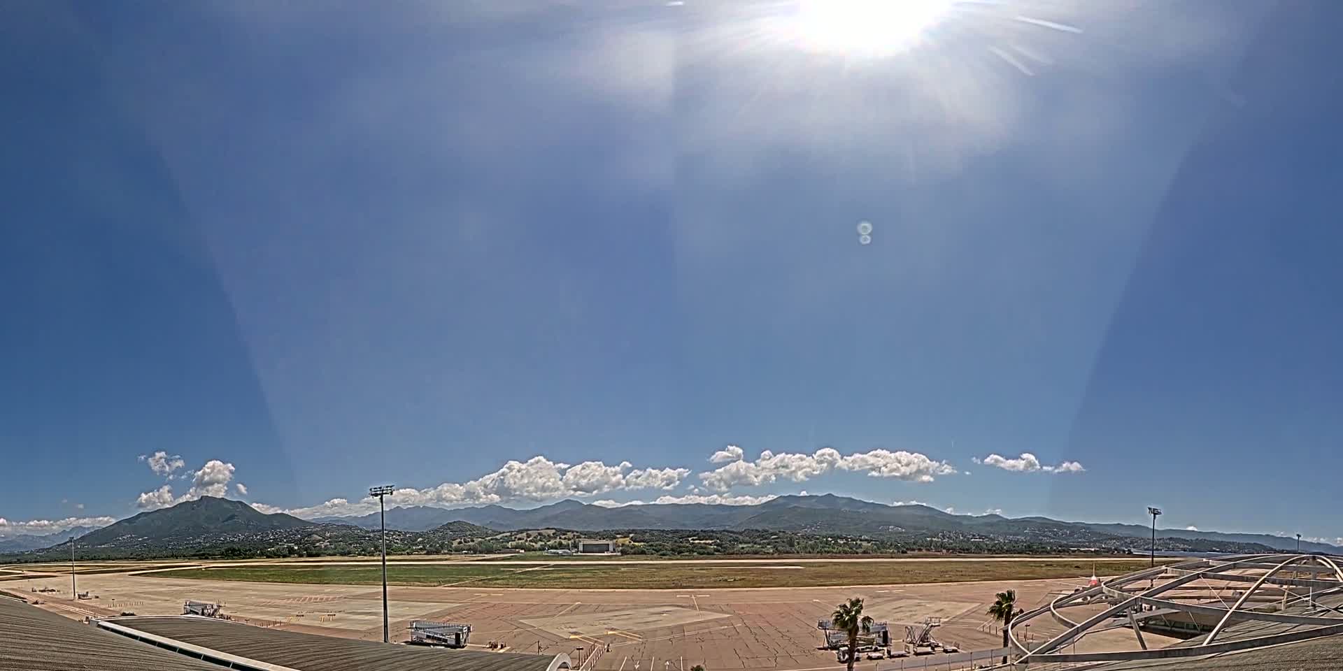 A sunny day with partly cloudy skies overlooks an airport tarmac and runway, with mountains in the distance.