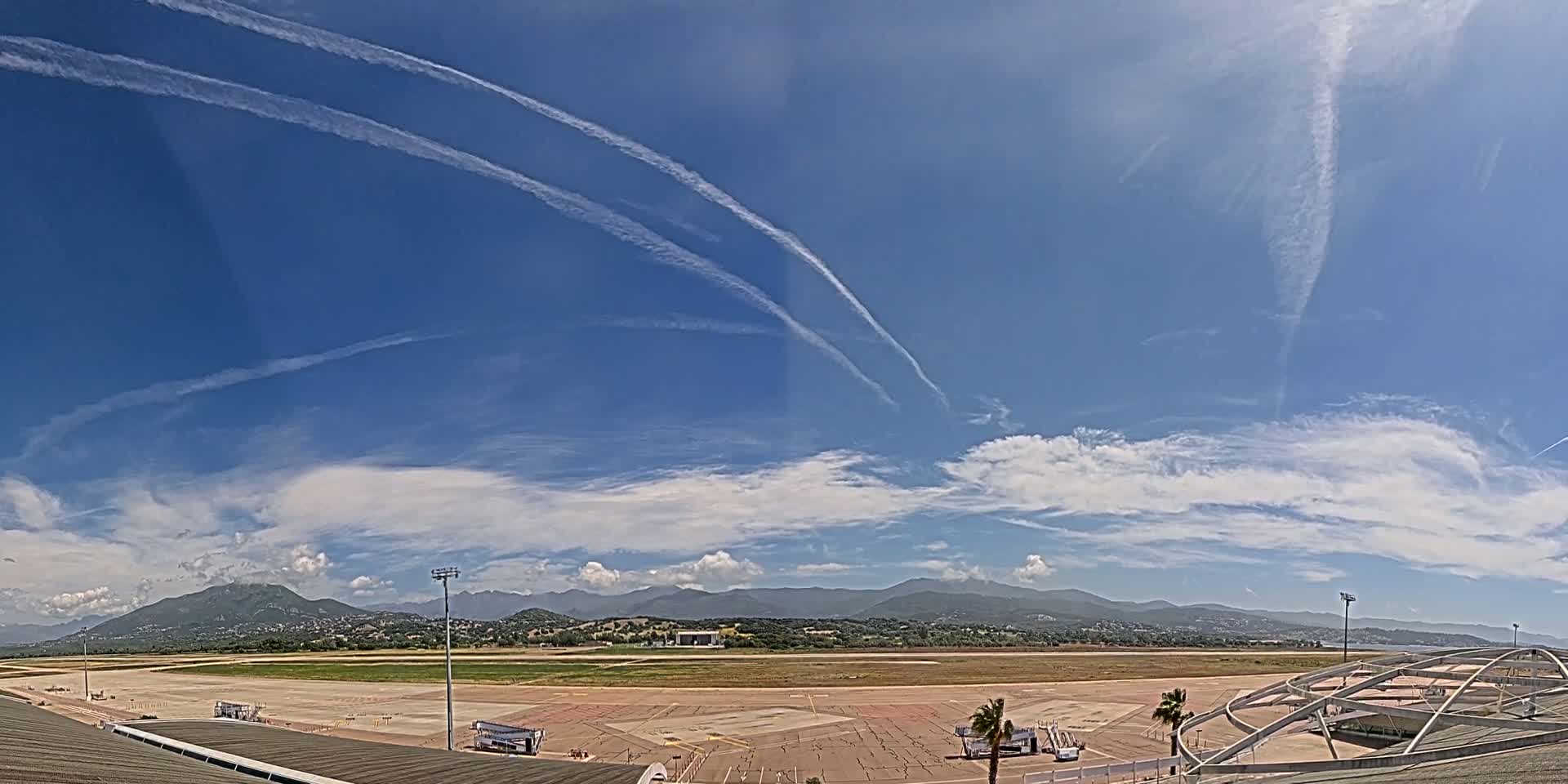 Under a mostly sunny sky with contrails and scattered cumulus clouds, an airport tarmac is visible, with mountains in the distance.