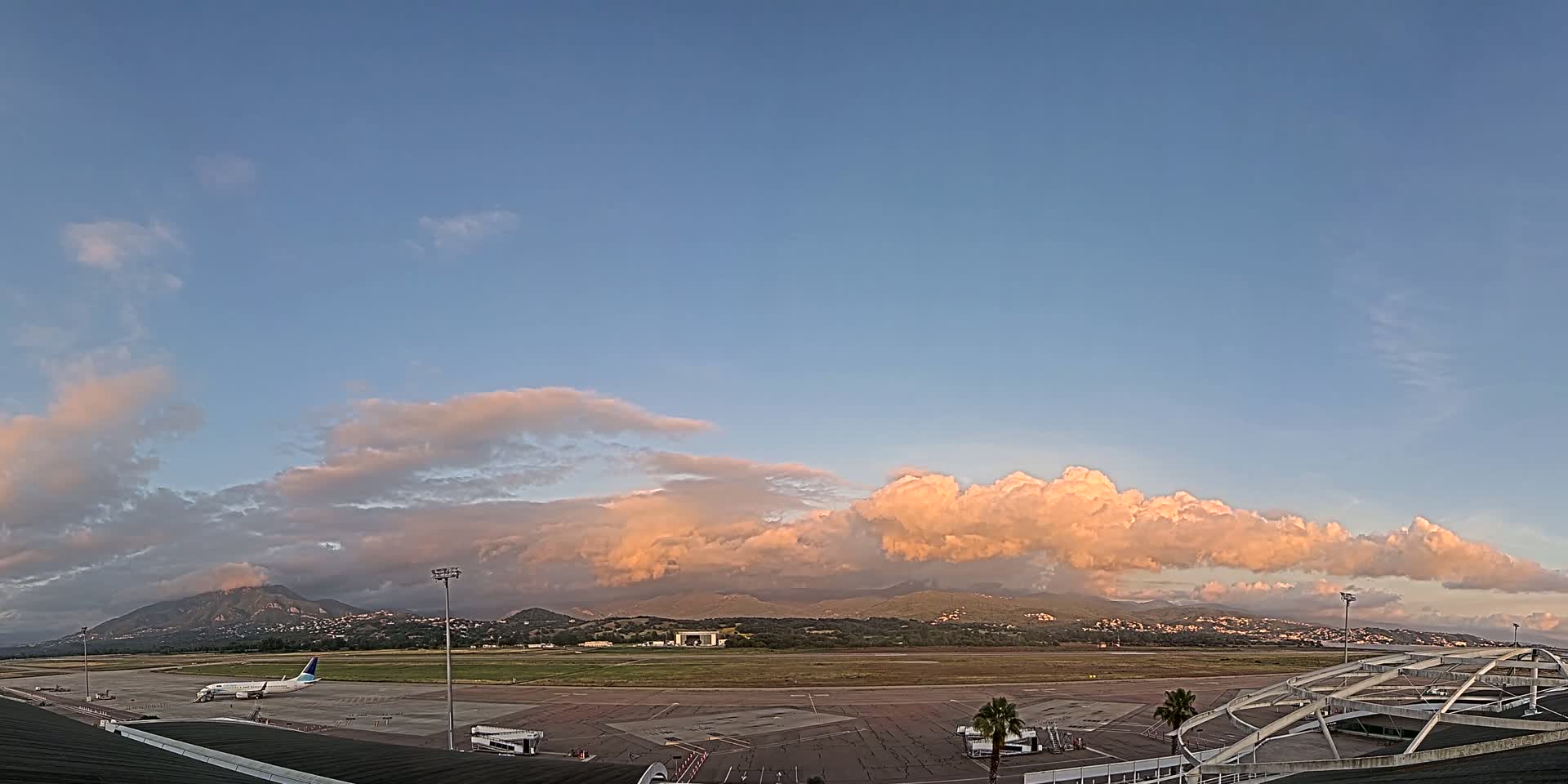 A passenger airplane sits on an airport tarmac under a partly cloudy sky with pink-tinged clouds at sunset, mountains visible in the distance.