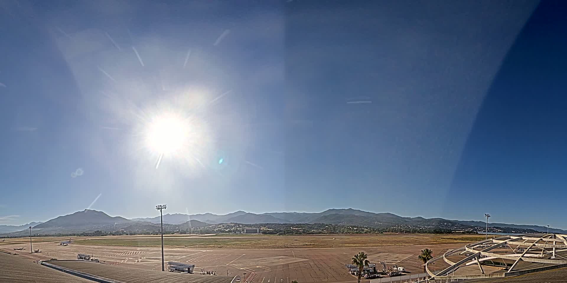 A sunny day at an airport shows a large tarmac with several service vehicles, a runway extending to distant mountains under a clear blue sky.