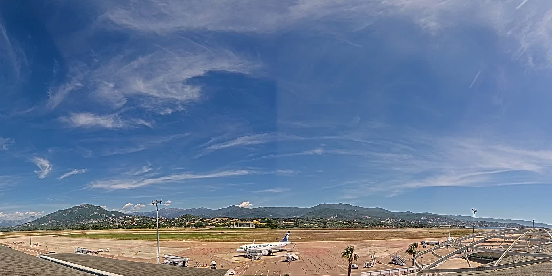 A passenger plane sits on the tarmac of an airport under a mostly clear blue sky with wispy clouds, mountains visible in the distance.