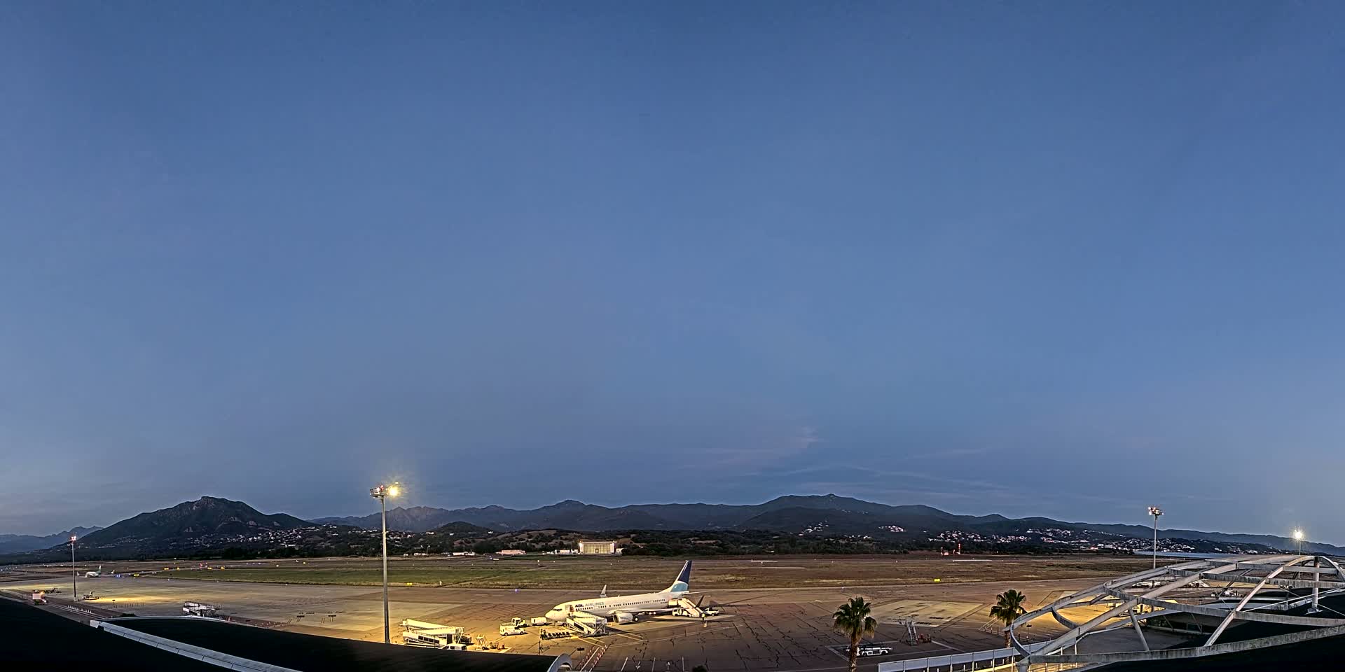 A passenger jet is parked at an airport tarmac at dusk, with mountains visible in the distance under a clear blue sky.