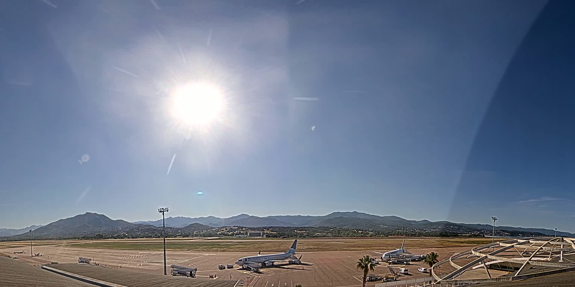 Two airplanes are parked on a sunny airport tarmac with mountains visible in the distance.