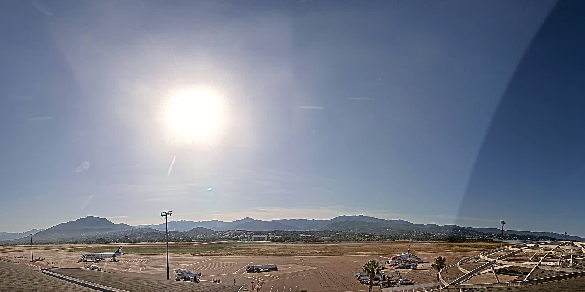 A sunny day at an airport shows several planes on the tarmac with mountains in the background.