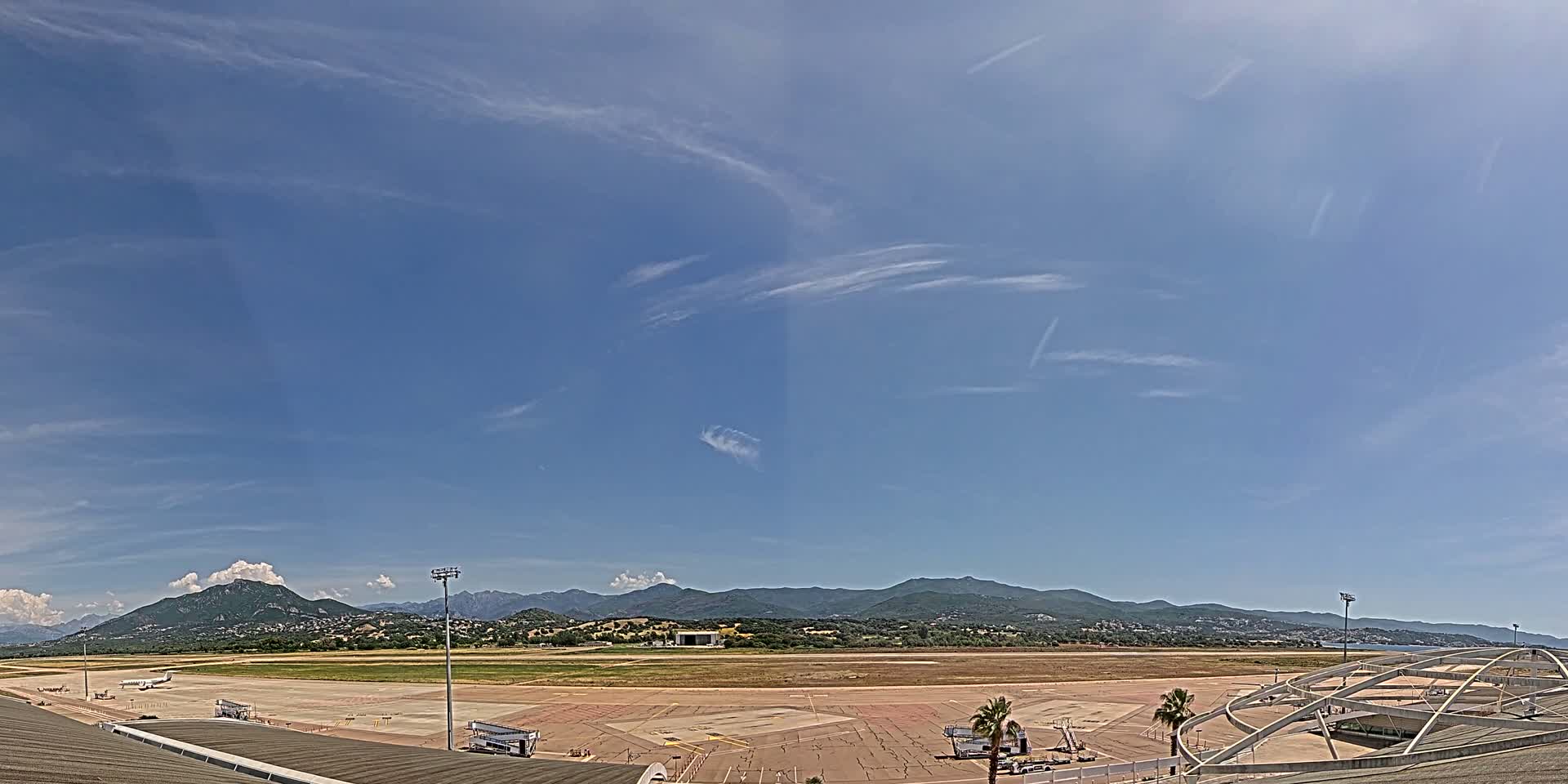 An airport tarmac with a few planes and jet bridges sits beneath a mostly clear blue sky with wispy cirrus clouds and a mountain range in the distance.