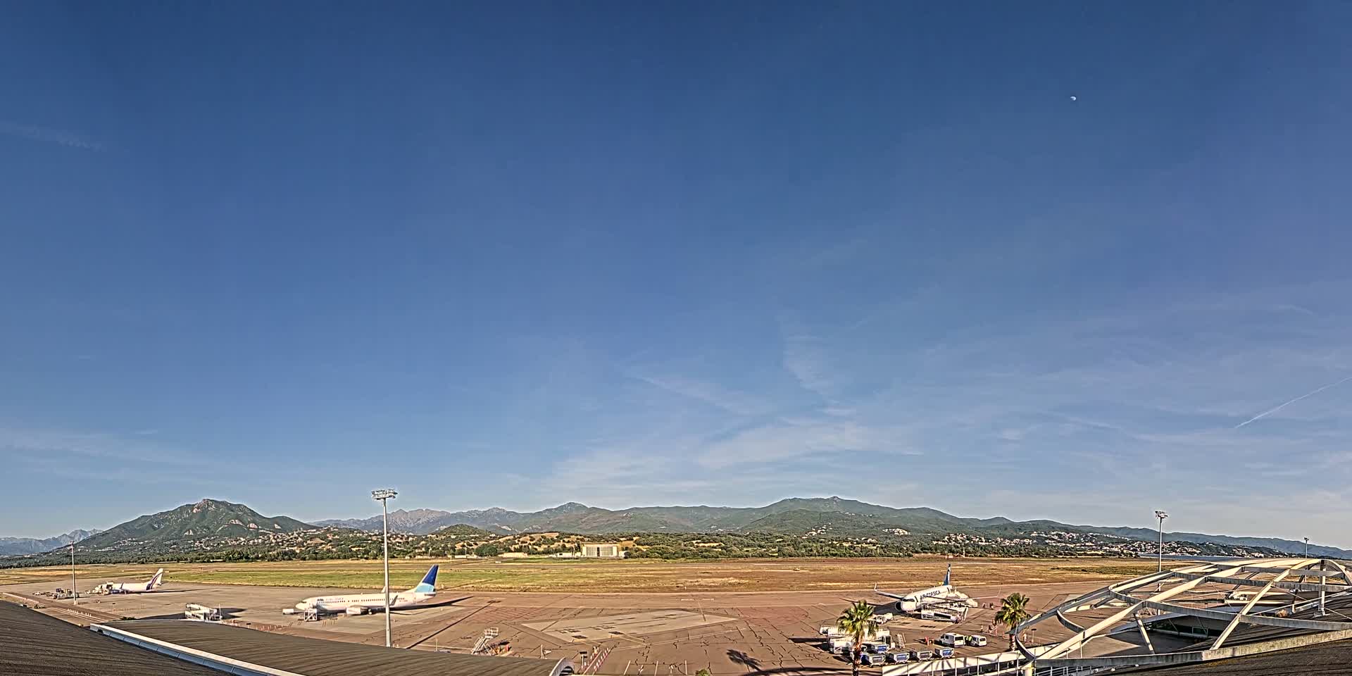 Under a clear blue sky, several airplanes are parked on an airport tarmac with mountains visible in the distance.