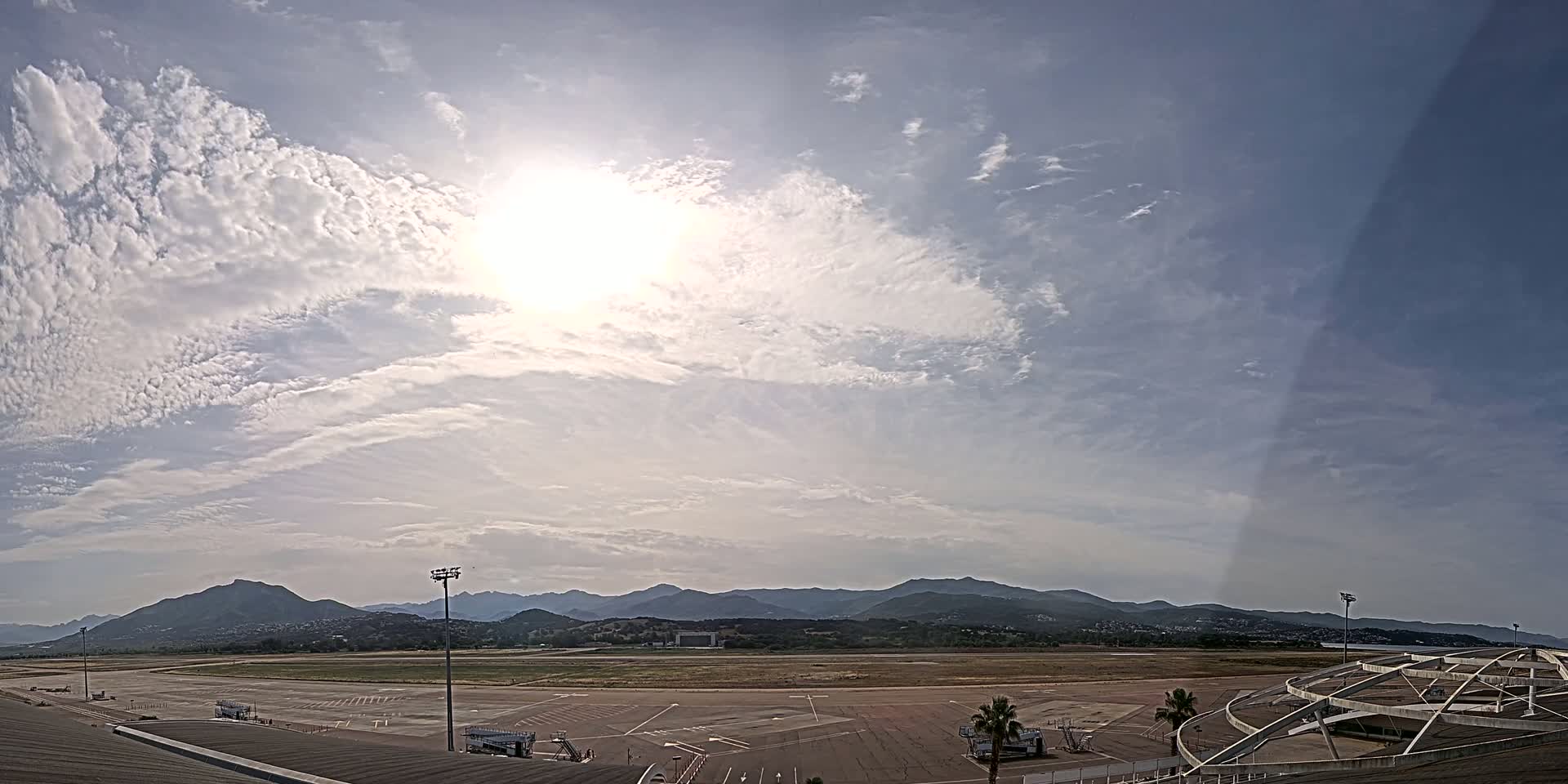 A mostly sunny day with scattered clouds over a wide expanse of airport tarmac, backed by a range of distant mountains.