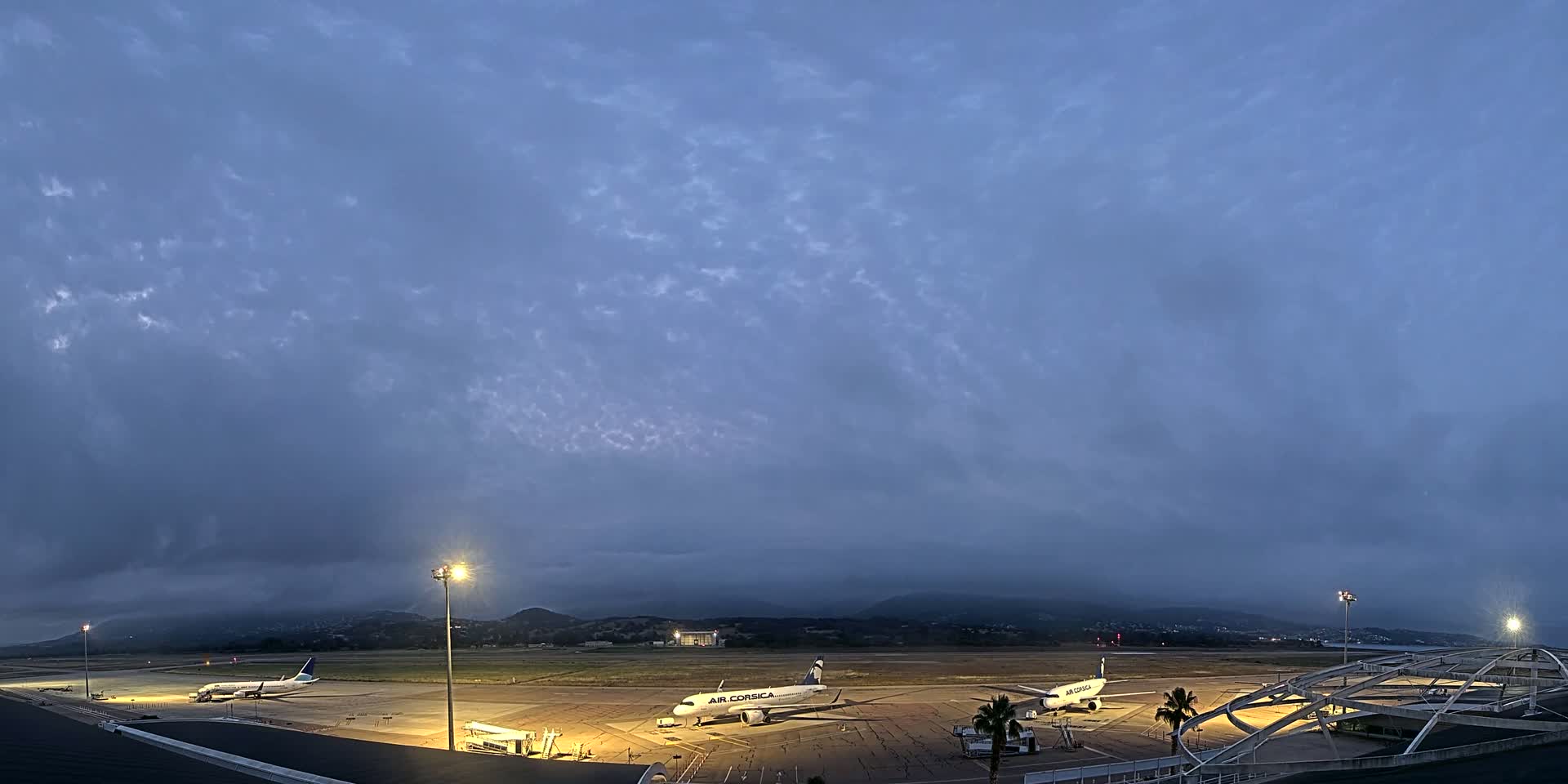 Several airplanes are parked on a dimly lit airport tarmac under a cloudy, twilight sky.