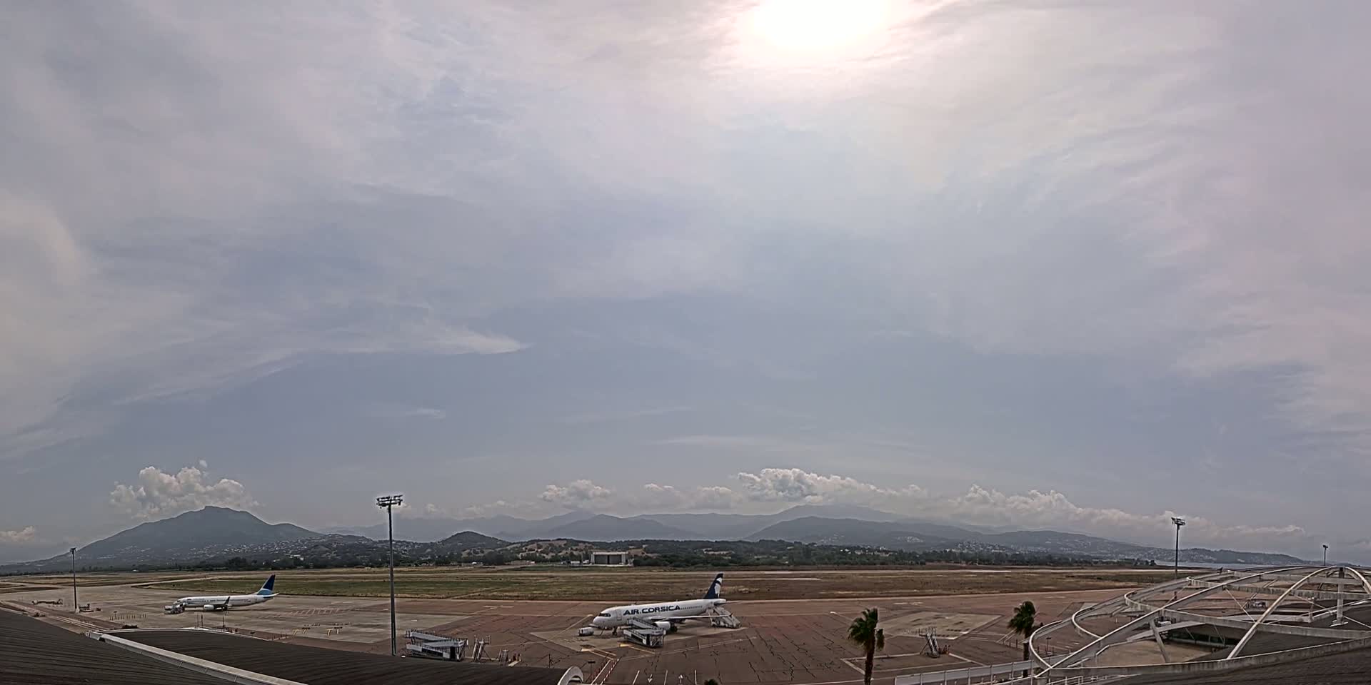 Two airplanes are parked on an airport tarmac under a partly cloudy sky, with mountains visible in the distance.