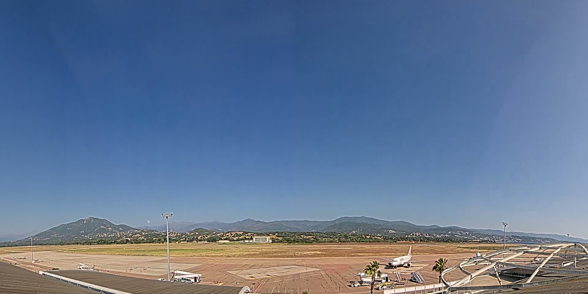 Under a clear blue sky, an airport tarmac shows a plane at a gate with mountains visible in the distance.