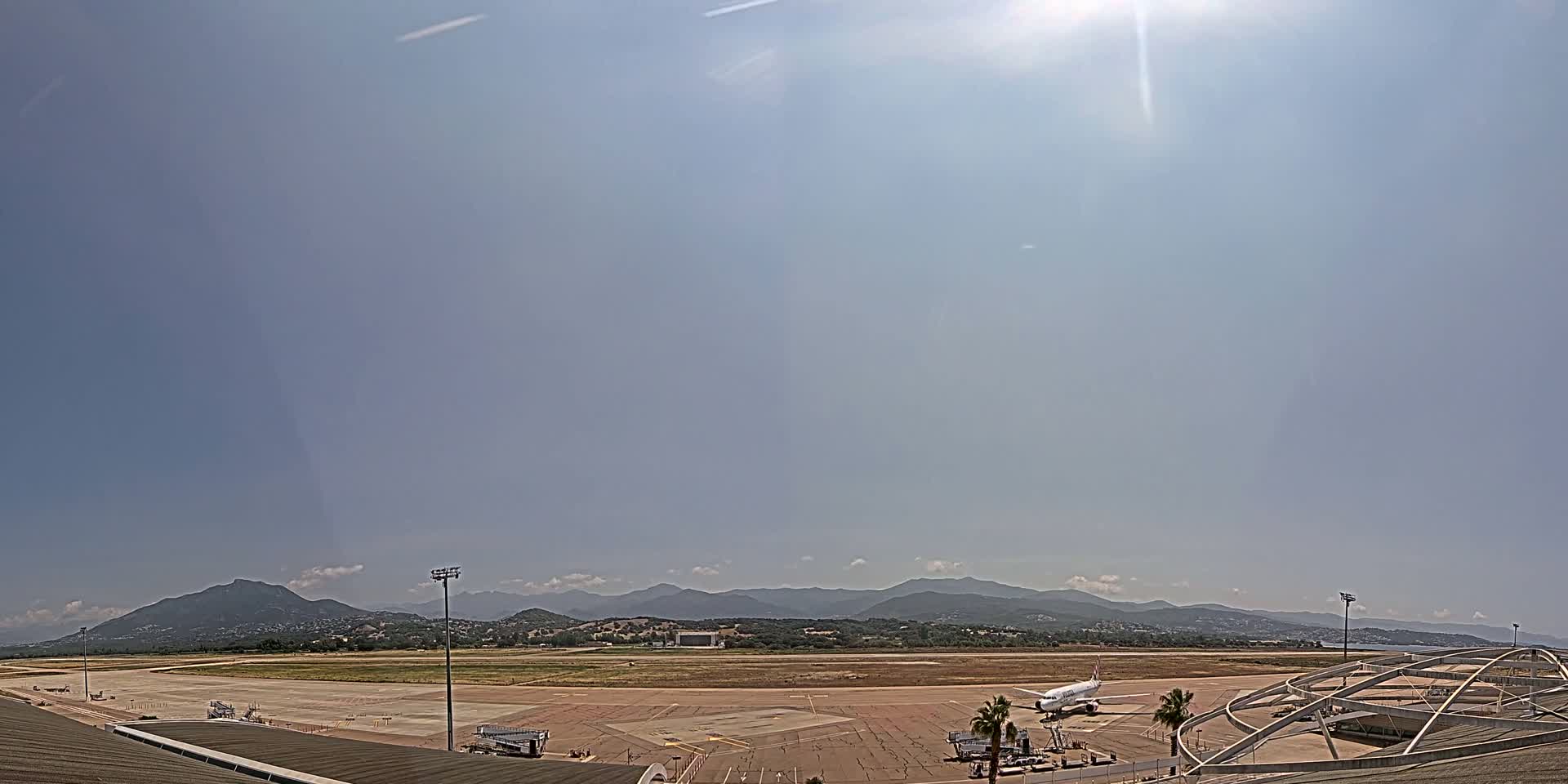 A sunny day reveals an airport tarmac with a plane parked near a terminal building, backed by a range of mountains in the distance.