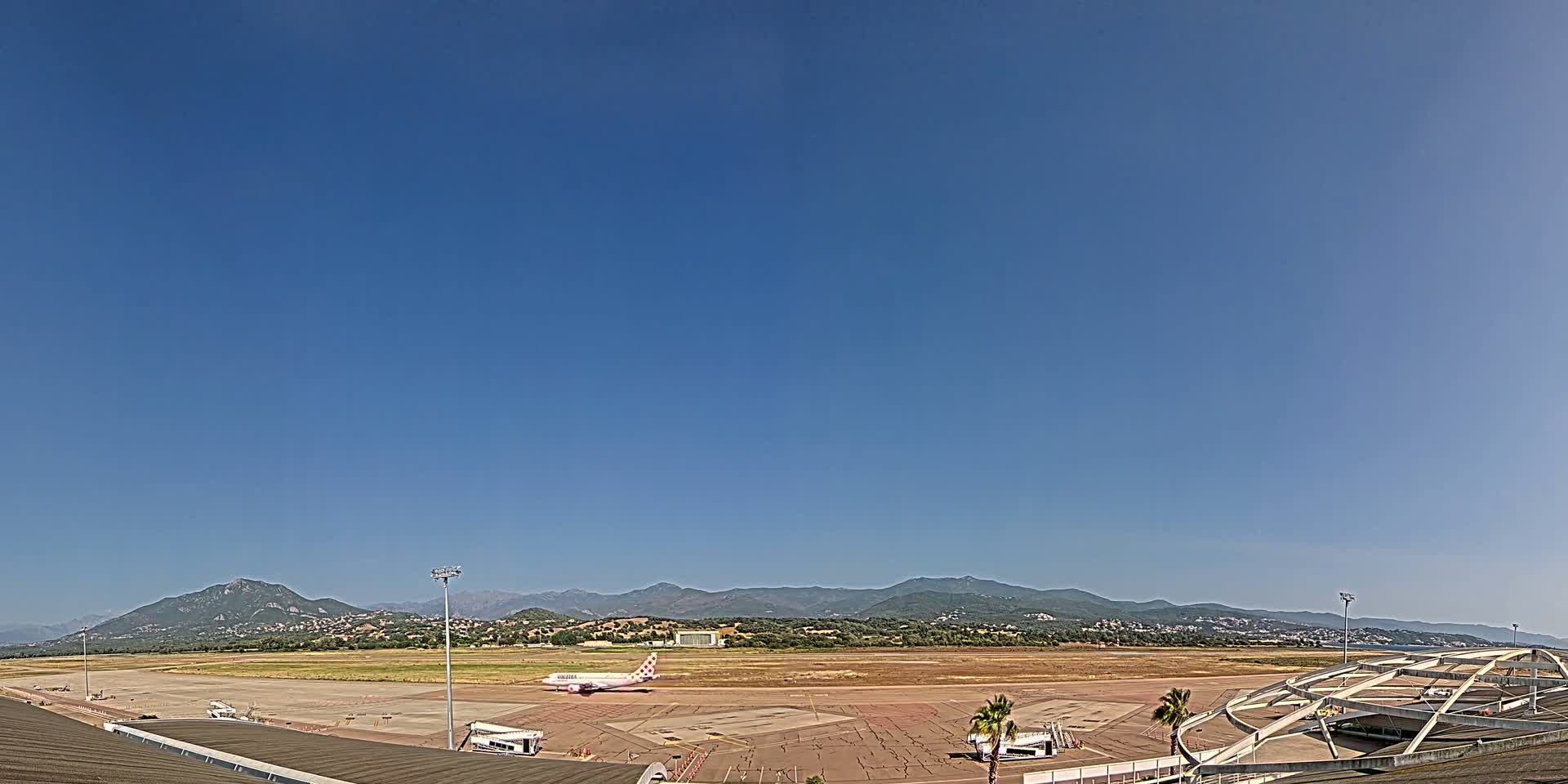 A pink-and-white airplane sits on a tarmac at an airport in front of distant mountains under a clear blue sky.