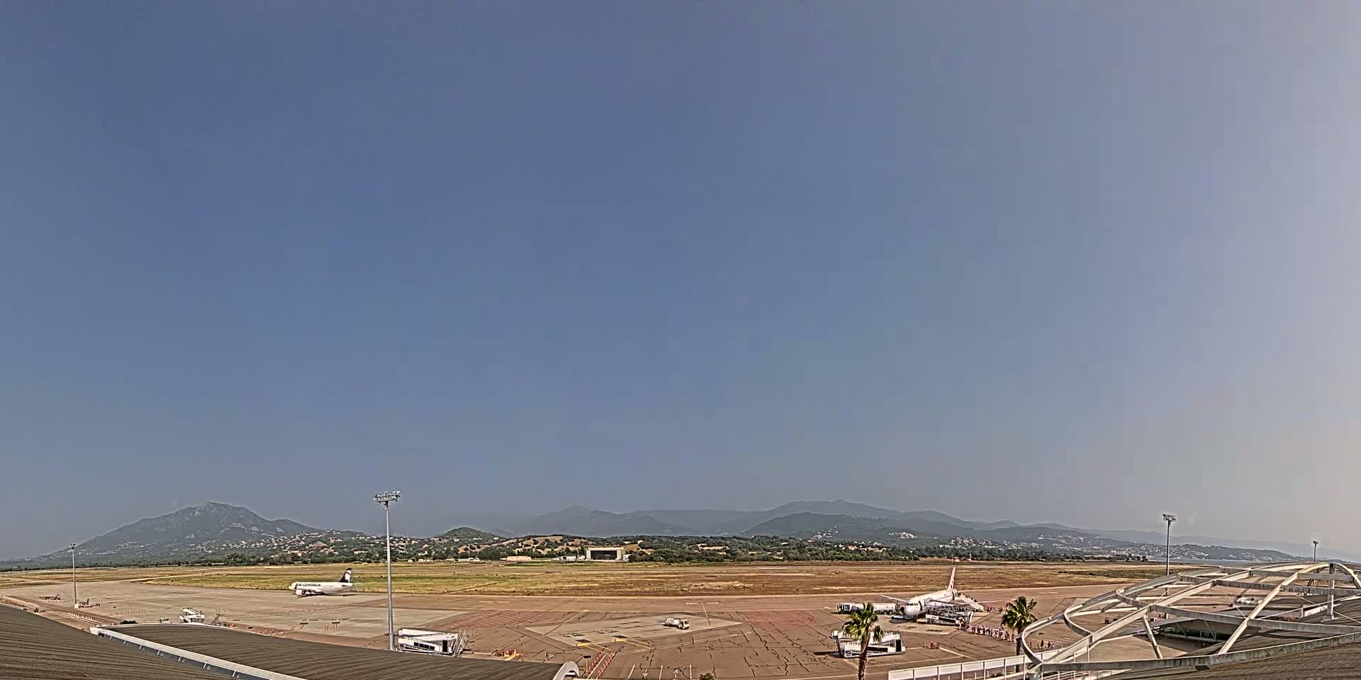 An airport tarmac with several airplanes, ground support equipment, and a portion of a terminal building is visible under a clear blue sky, with mountains in the background.