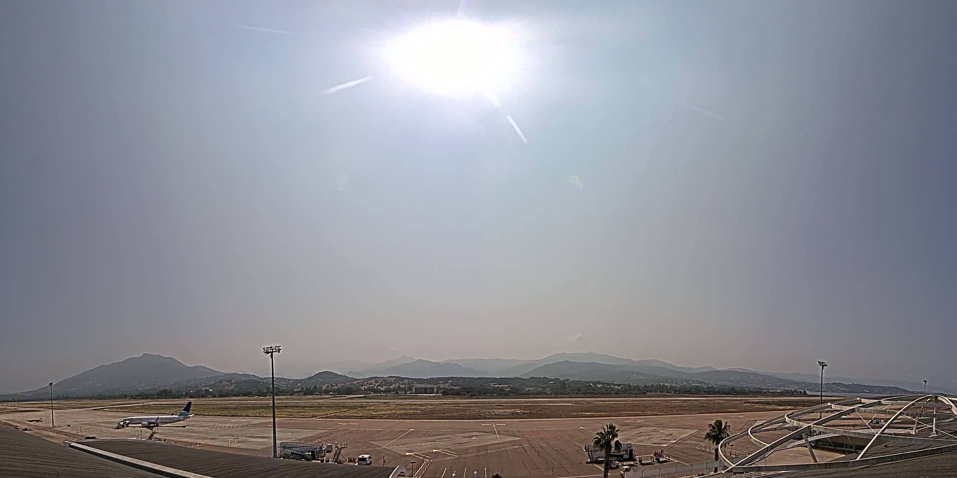 A sunny day at an airport shows a plane on the tarmac with mountains visible in the distance.