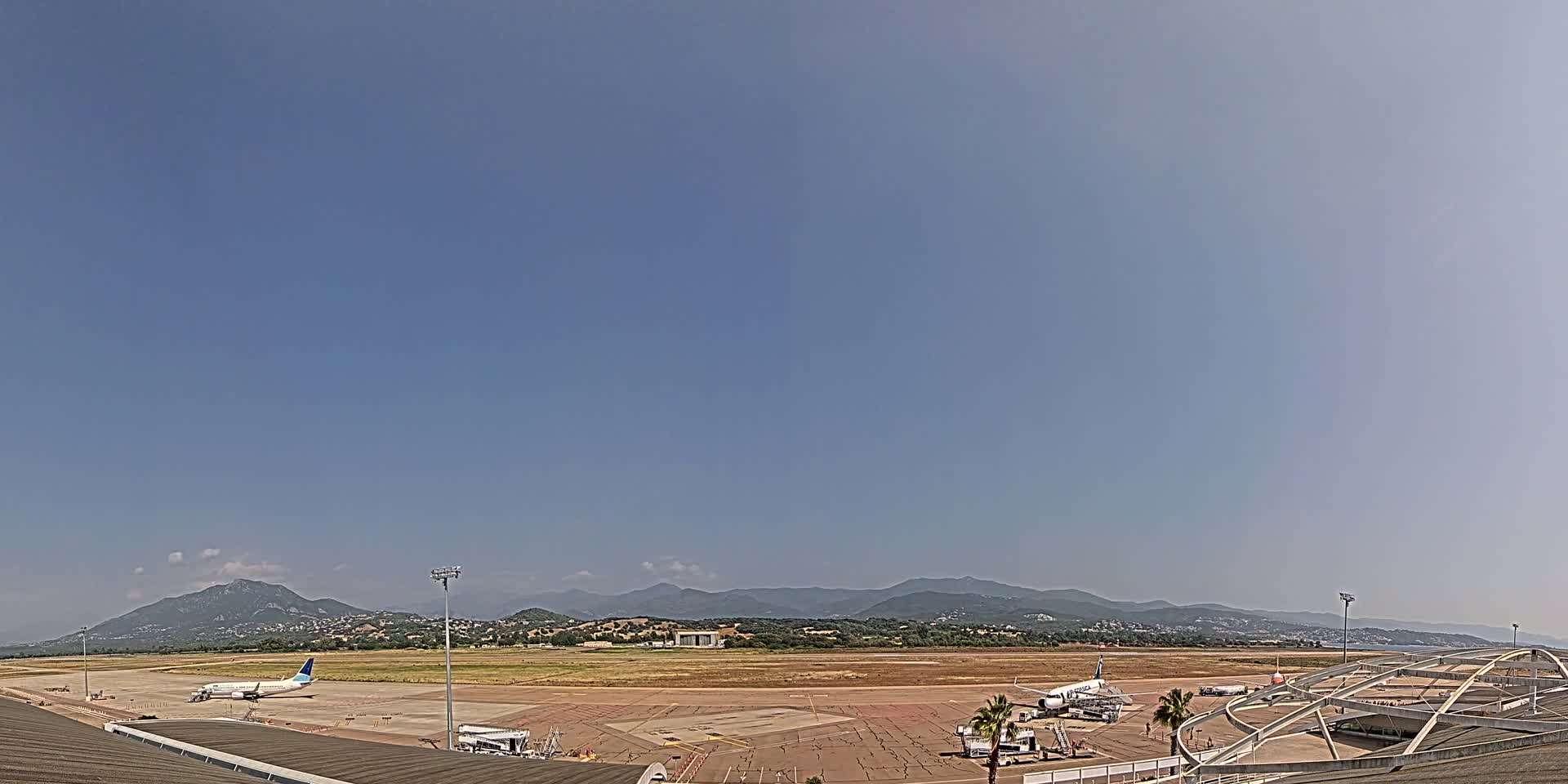 A mostly clear, sunny day at an airport with several airplanes parked on the tarmac, with mountains visible in the background.