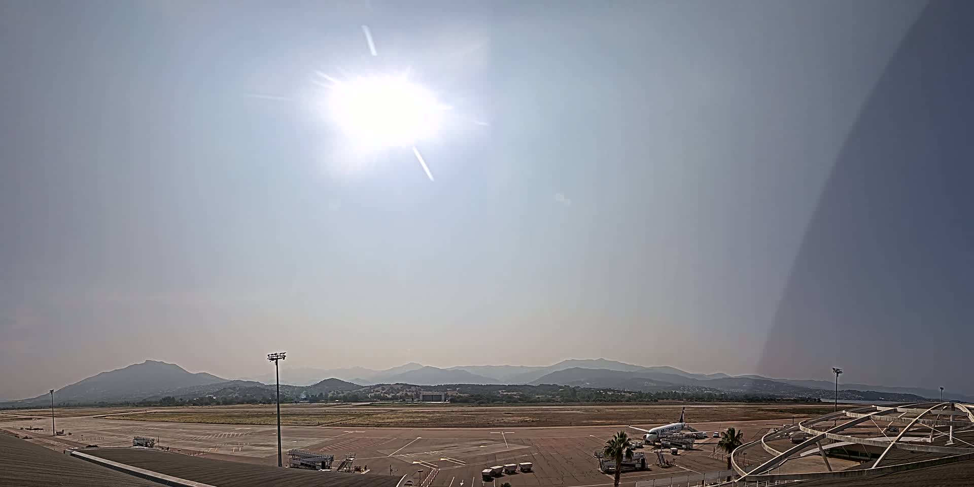 A sunny day at an airport with a plane parked on the tarmac, overlooking a range of distant mountains.