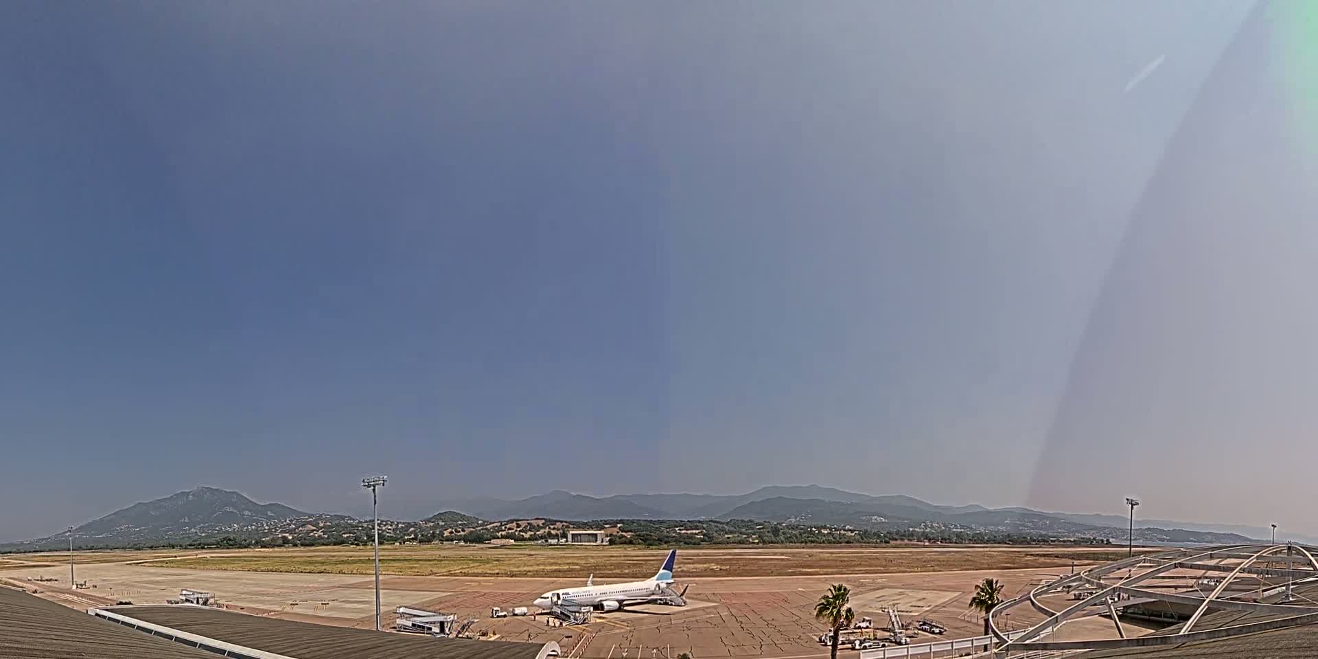 A passenger airplane sits on the tarmac of an airport on a clear, sunny day with mountains visible in the distance.