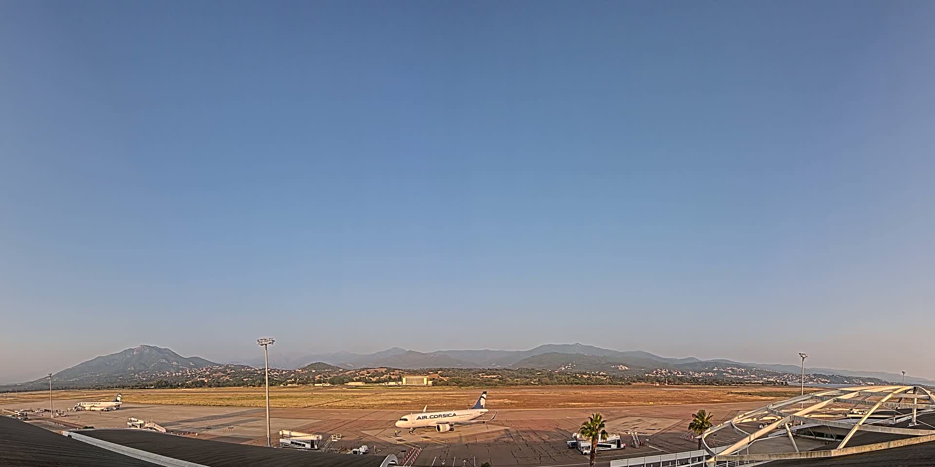 A passenger plane sits on a tarmac at an airport under a clear, blue sky with mountains in the background.
