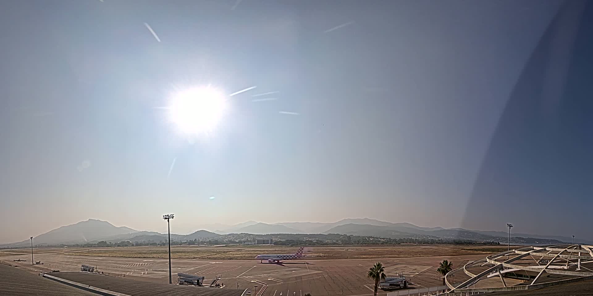 A bright, sunny day at an airport shows a plane on the tarmac with hazy mountains in the distance.