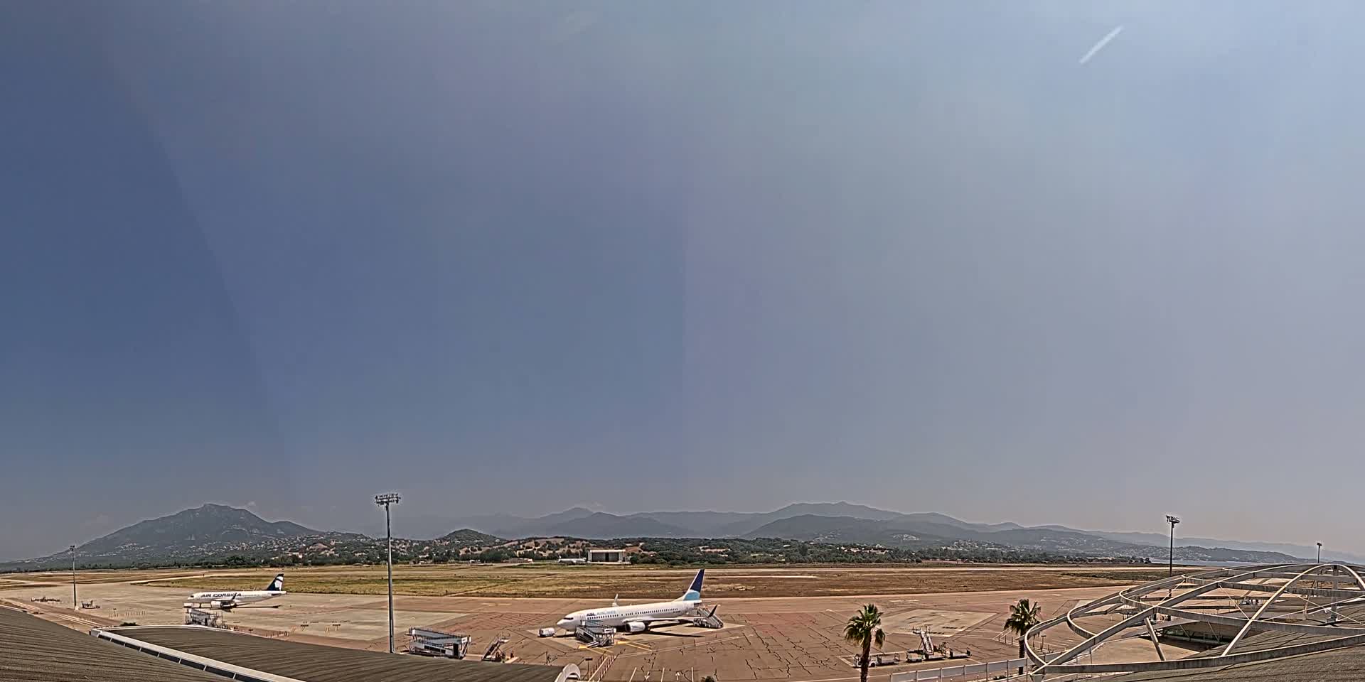 Two airplanes are parked on an airport tarmac under a clear, hazy sky, with mountains visible in the distance.