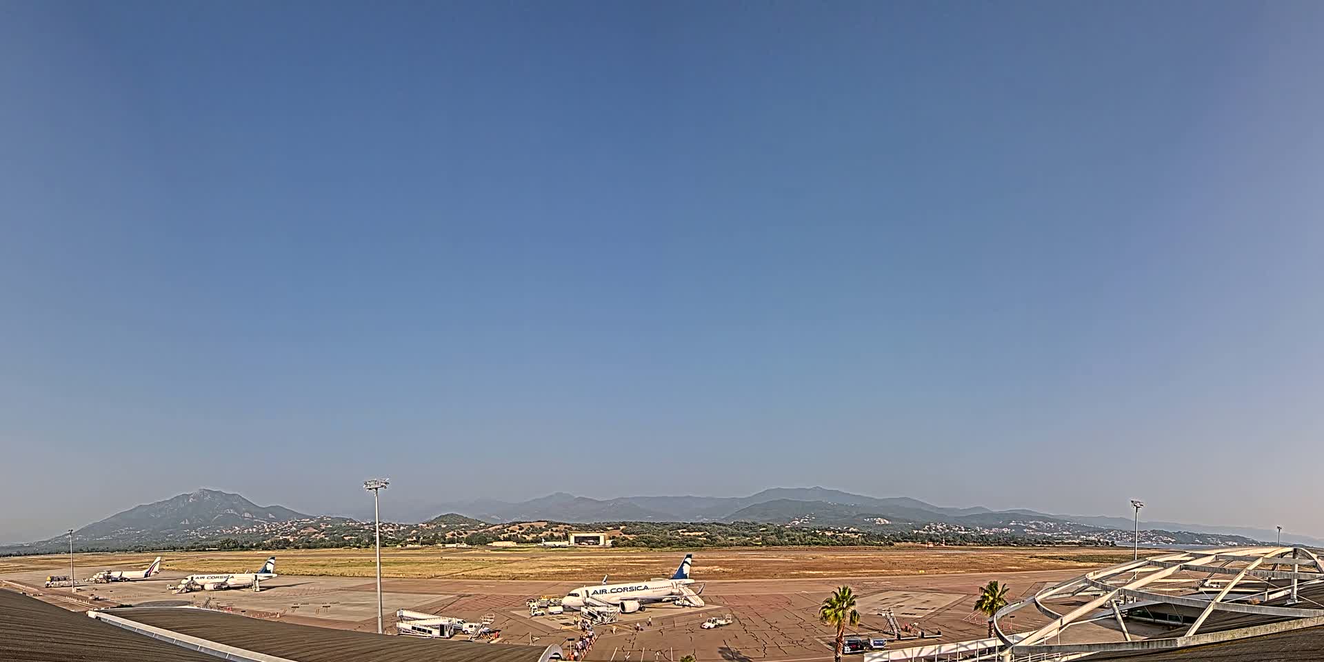 Several airplanes are parked on a tarmac at an airport on a clear, sunny day, with mountains visible in the distance.