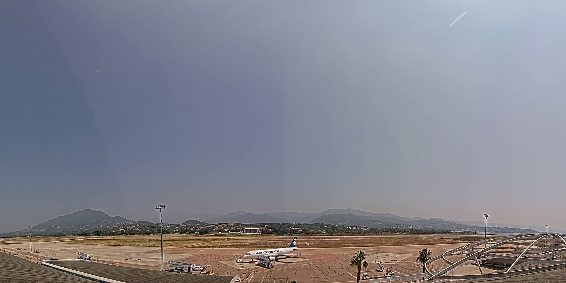 A passenger airplane sits on a tarmac at an airport on a clear, hazy day with mountains in the distance.