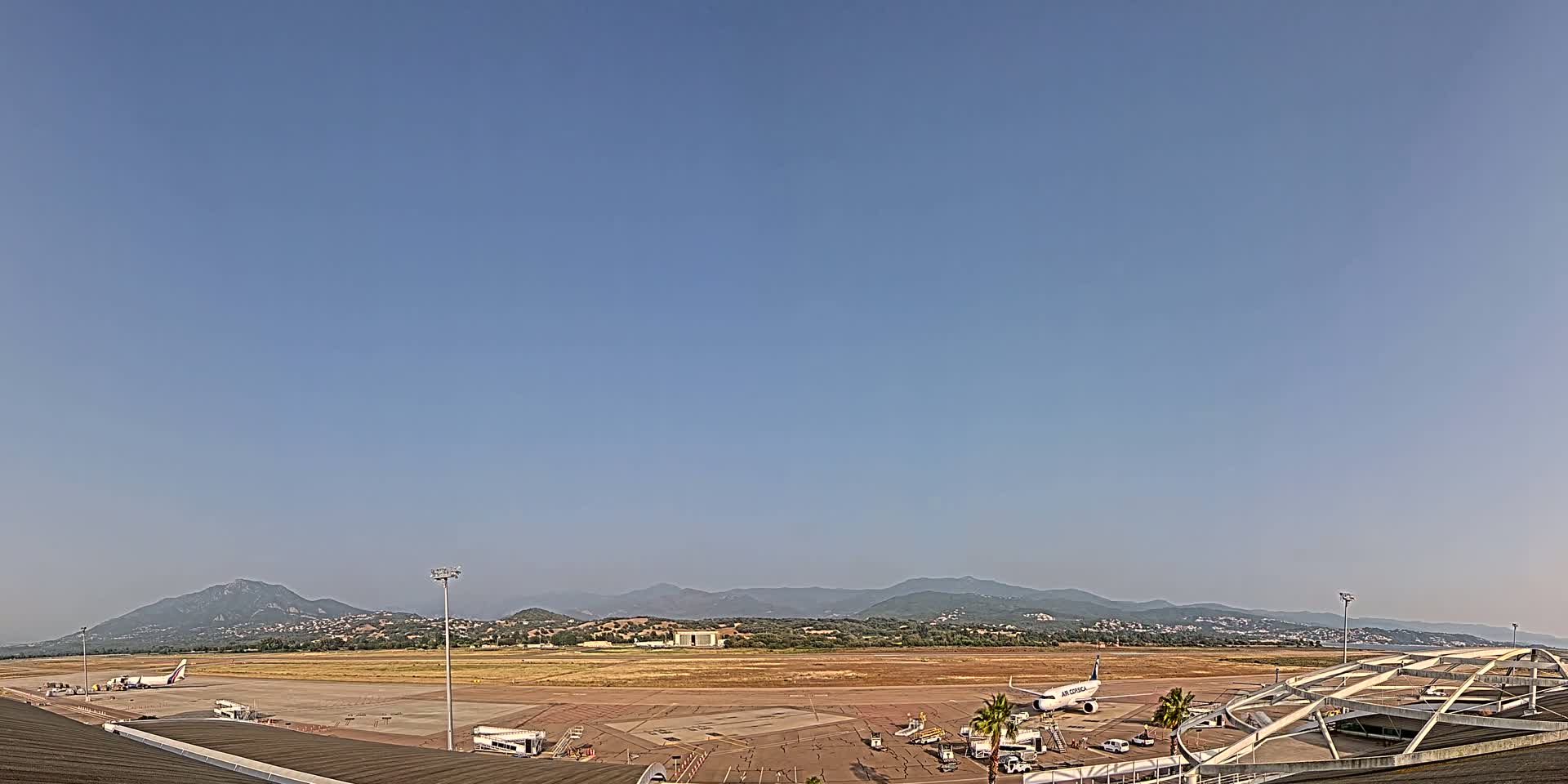 An airport tarmac with several airplanes and support vehicles, under a clear blue sky and backed by hazy mountains.
