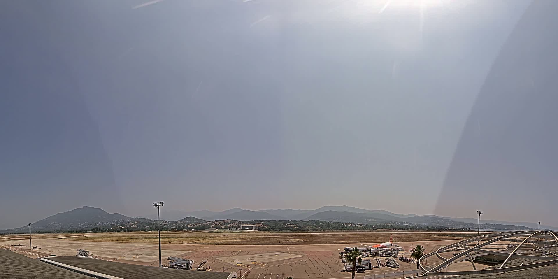 A mostly sunny, hazy day at an airport with a runway and several aircraft visible, set against a backdrop of distant mountains.