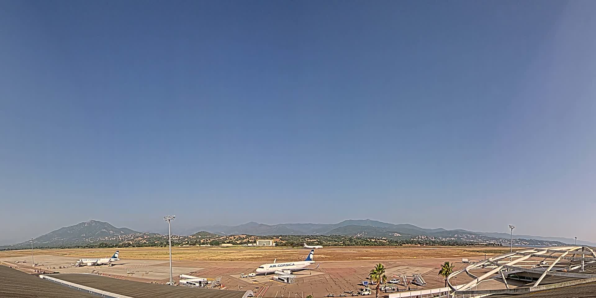 Under a clear blue sky, several airplanes are parked on a tarmac at an airport, with mountains visible in the distance.