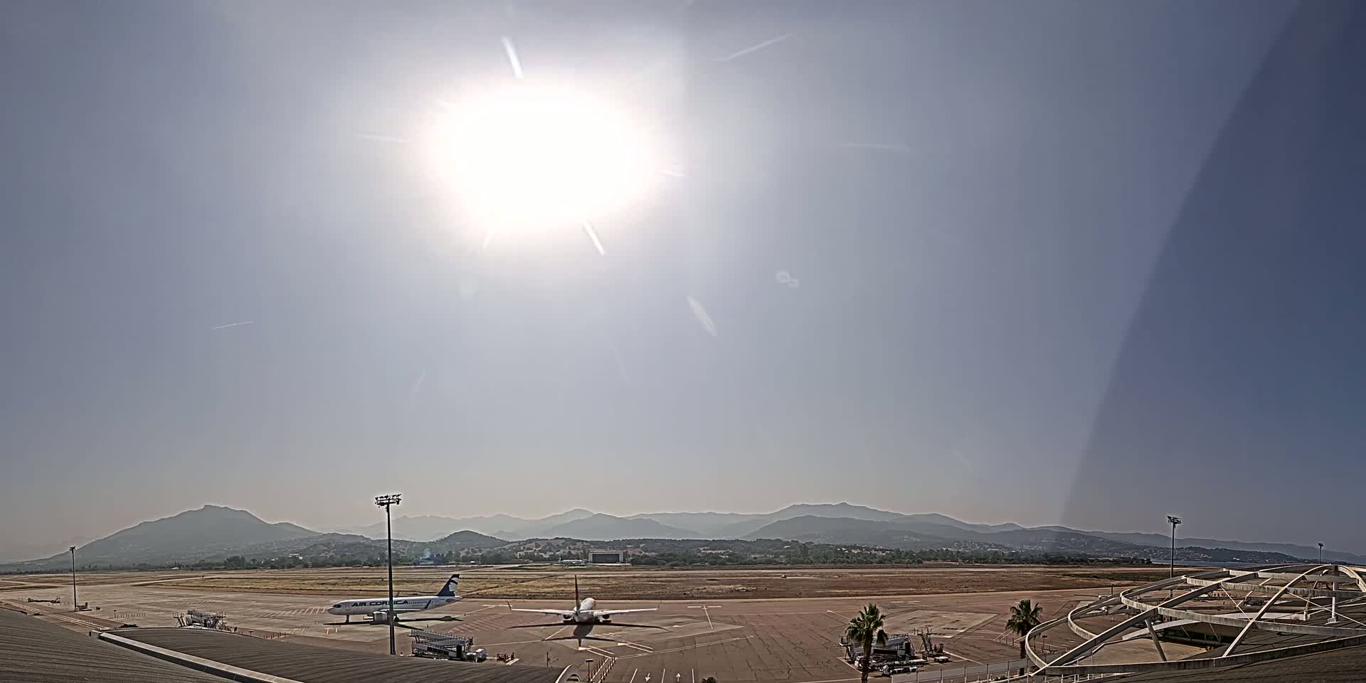 A bright, sunny day at an airport with a plane on the tarmac and mountains in the background.