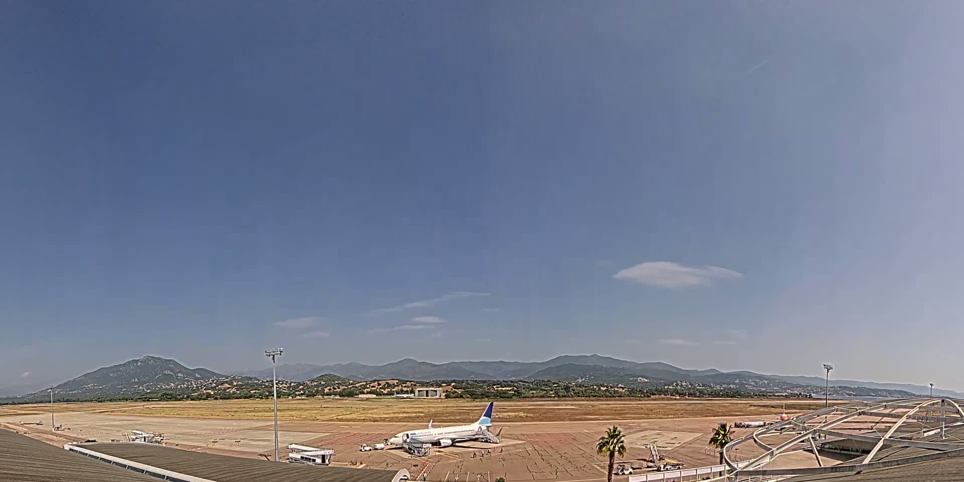 A passenger plane sits on a tarmac at an airport on a mostly clear day, with mountains visible in the distance.
