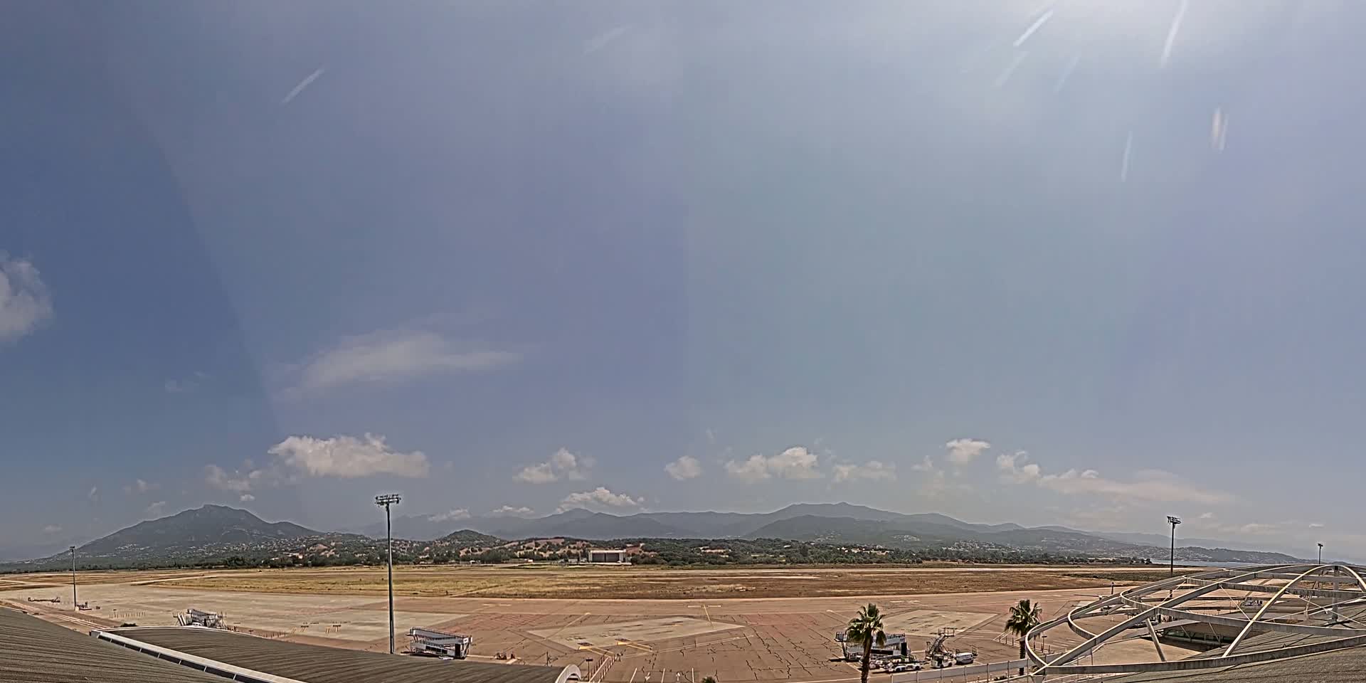 A mostly sunny day reveals an airport tarmac, a few service vehicles, and distant mountains under a pale blue sky with scattered clouds.