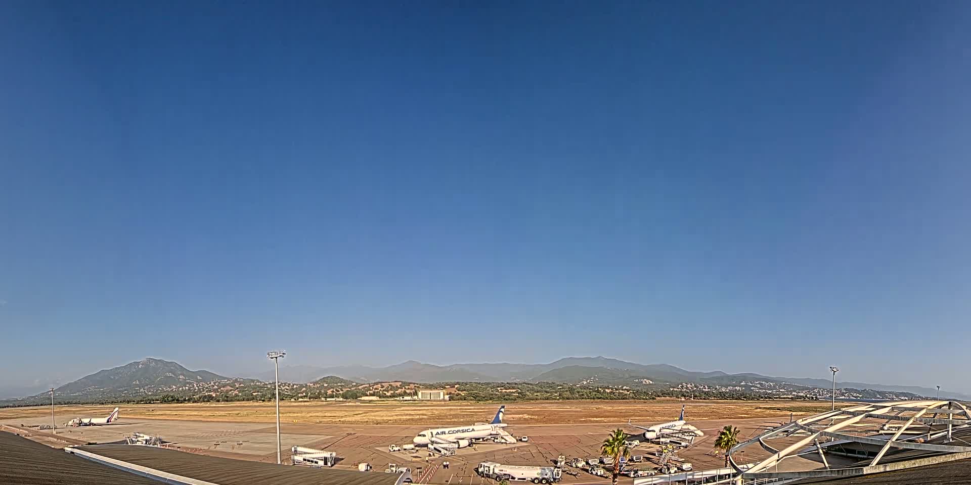 Several airplanes are parked on a tarmac at an airport, with mountains visible in the distance under a clear blue sky.