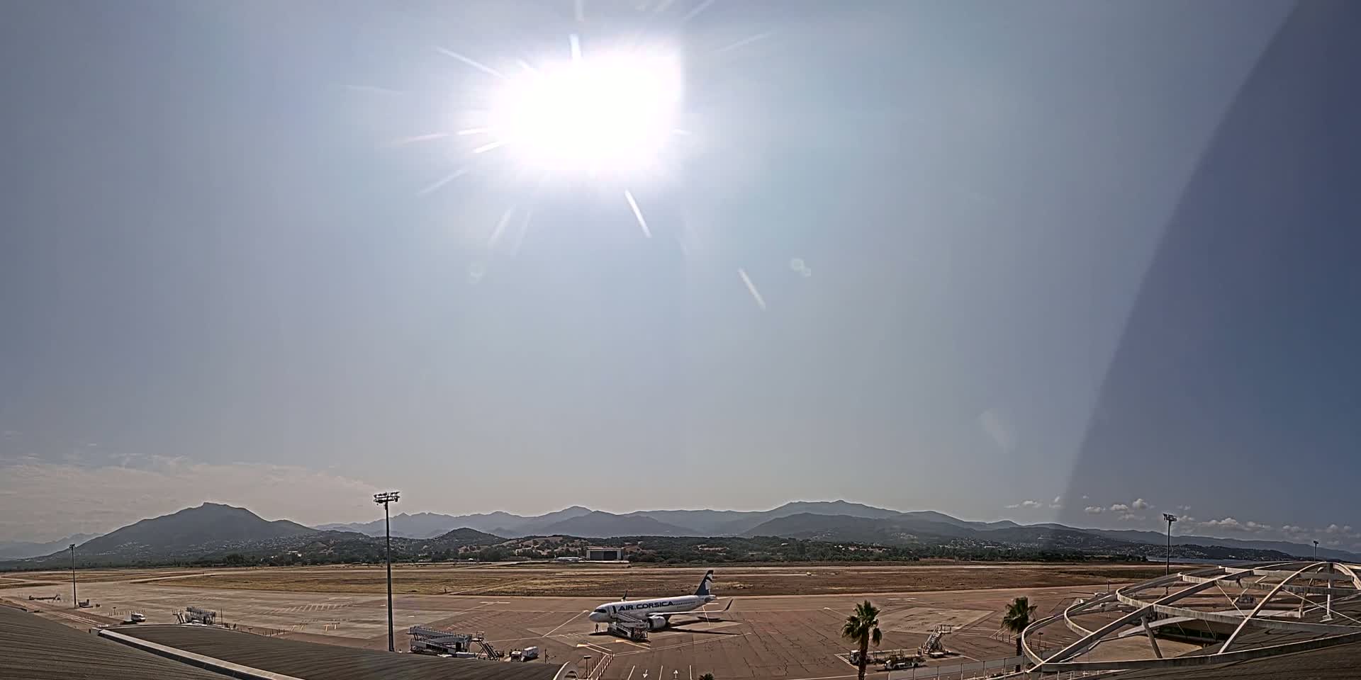A passenger airplane sits on a tarmac at an airport on a sunny day, with mountains visible in the distance.