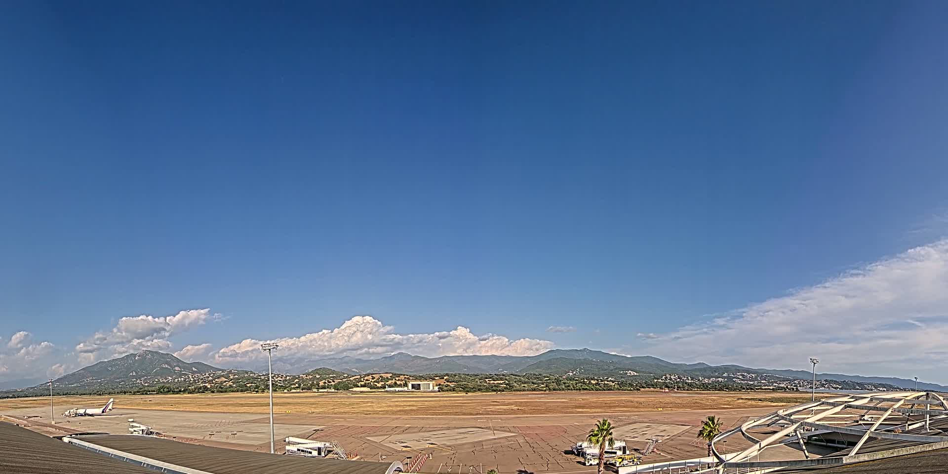 A mostly clear blue sky with some cumulus clouds over a tarmac airport with a single airplane parked near a terminal building and mountains in the distance.