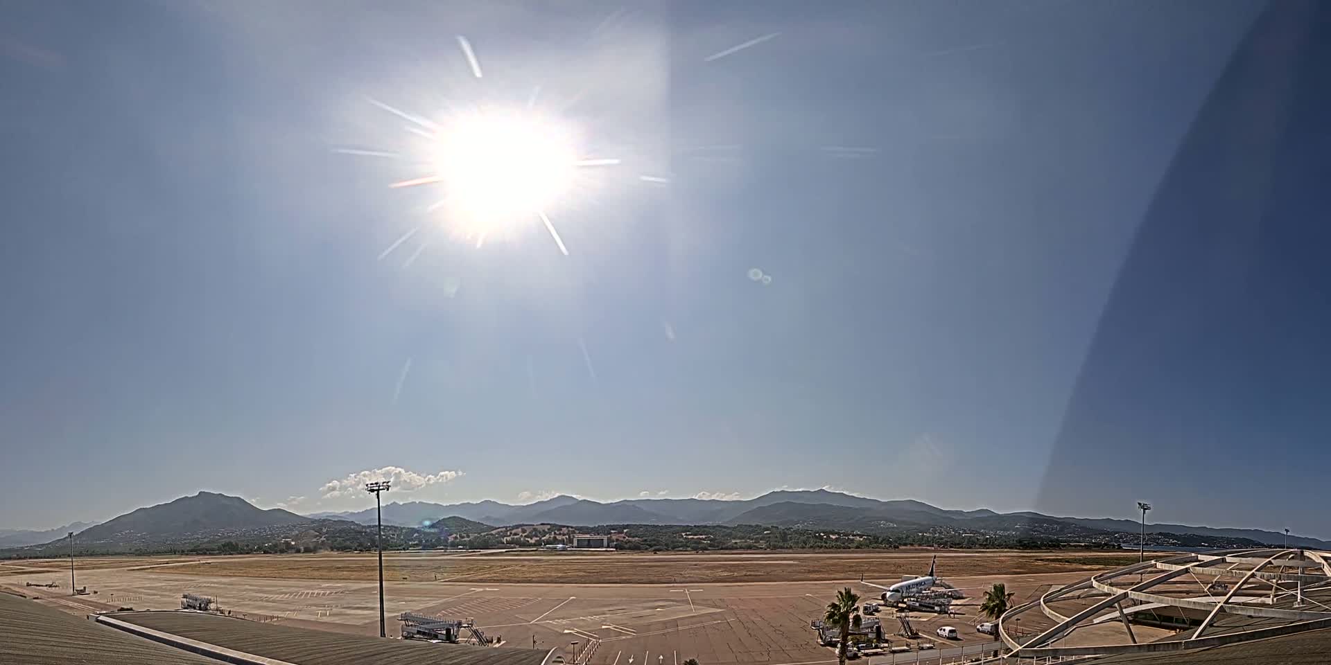 A bright, sunny day shows a mostly empty airport tarmac with a single airplane and mountains in the distance.
