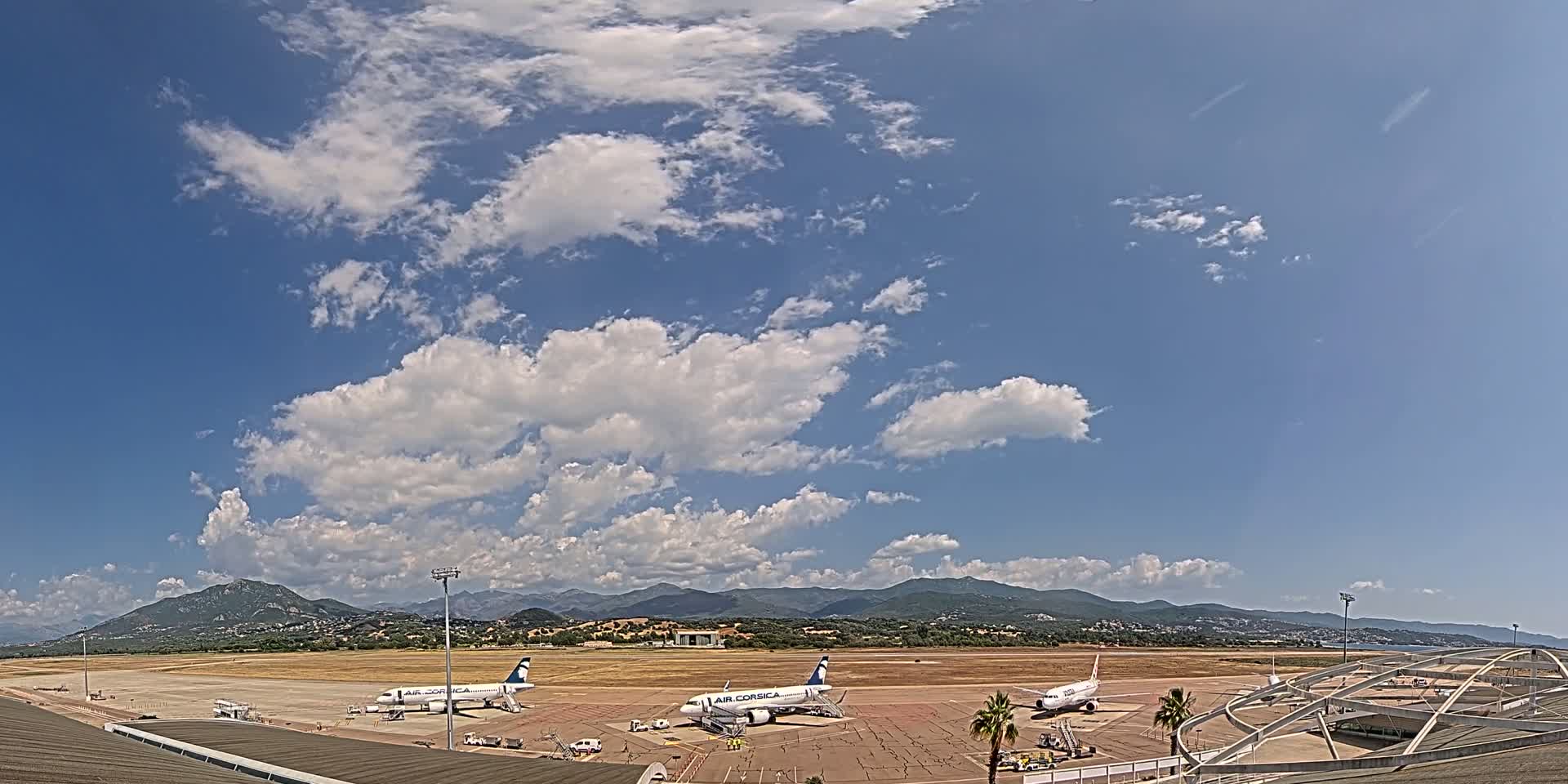 Three airplanes are parked on an airport tarmac under a partly cloudy sky, with mountains visible in the distance.