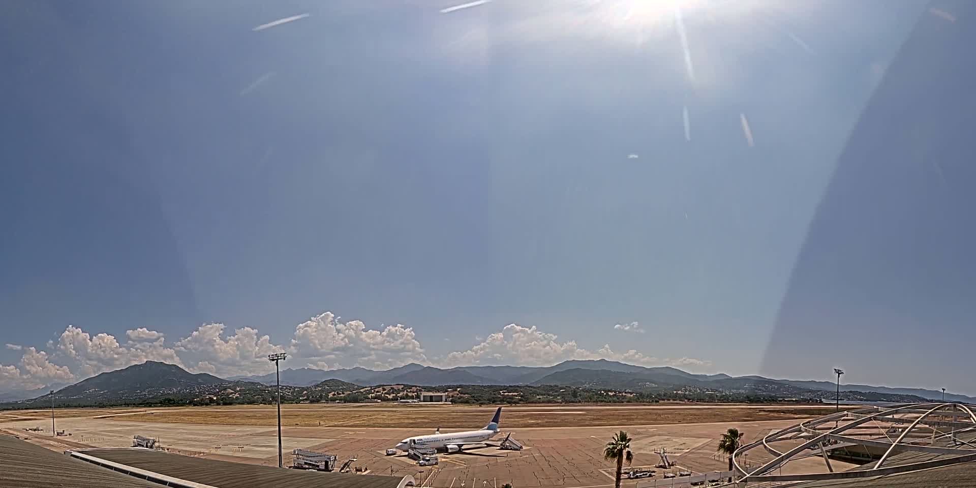 A passenger airplane sits on a tarmac at an airport under a mostly sunny sky with some cumulus clouds and mountains in the distance.