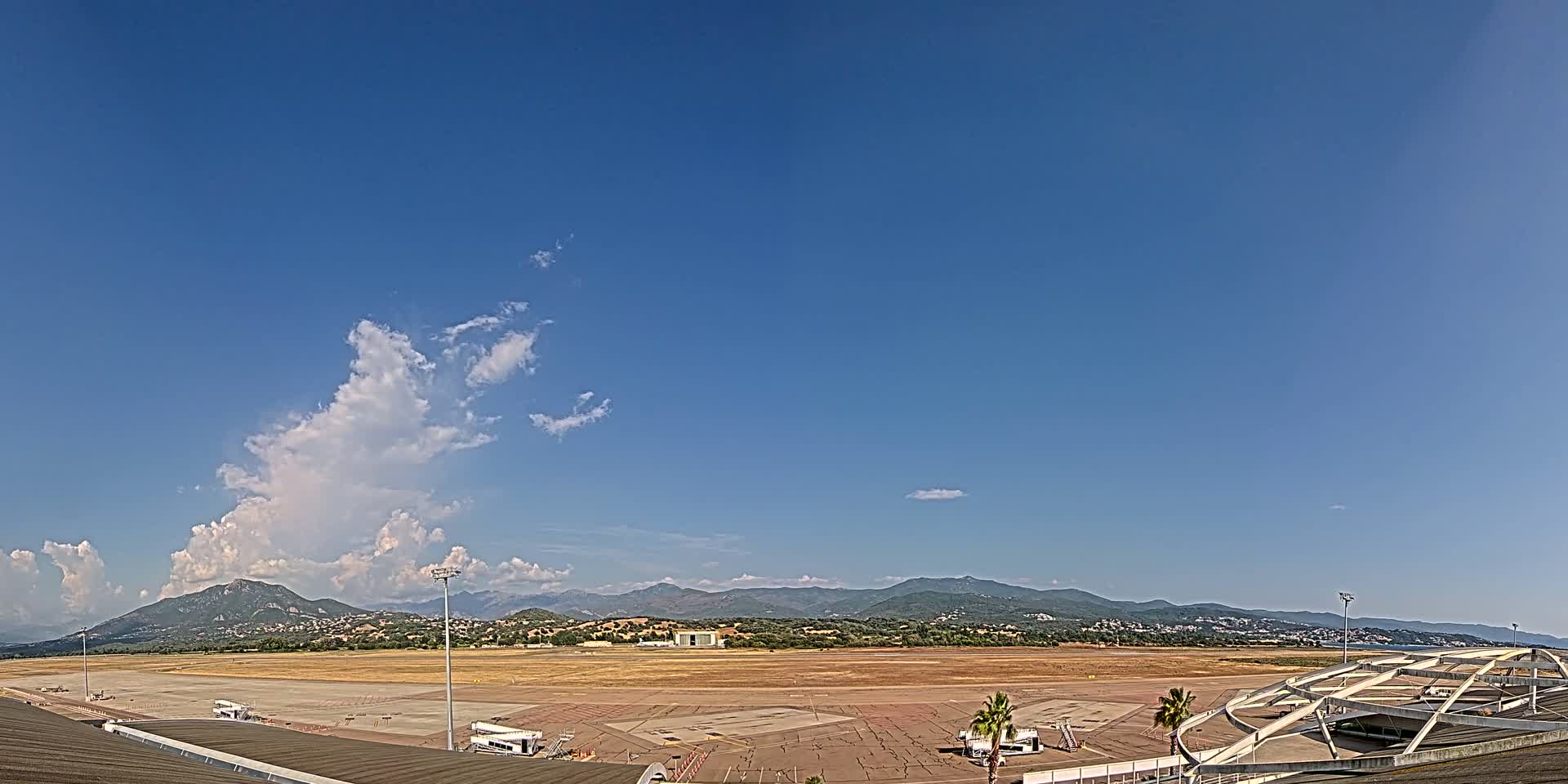 An airport tarmac with a few service vehicles is visible, backed by a range of mountains under a mostly clear blue sky with some cumulus clouds.