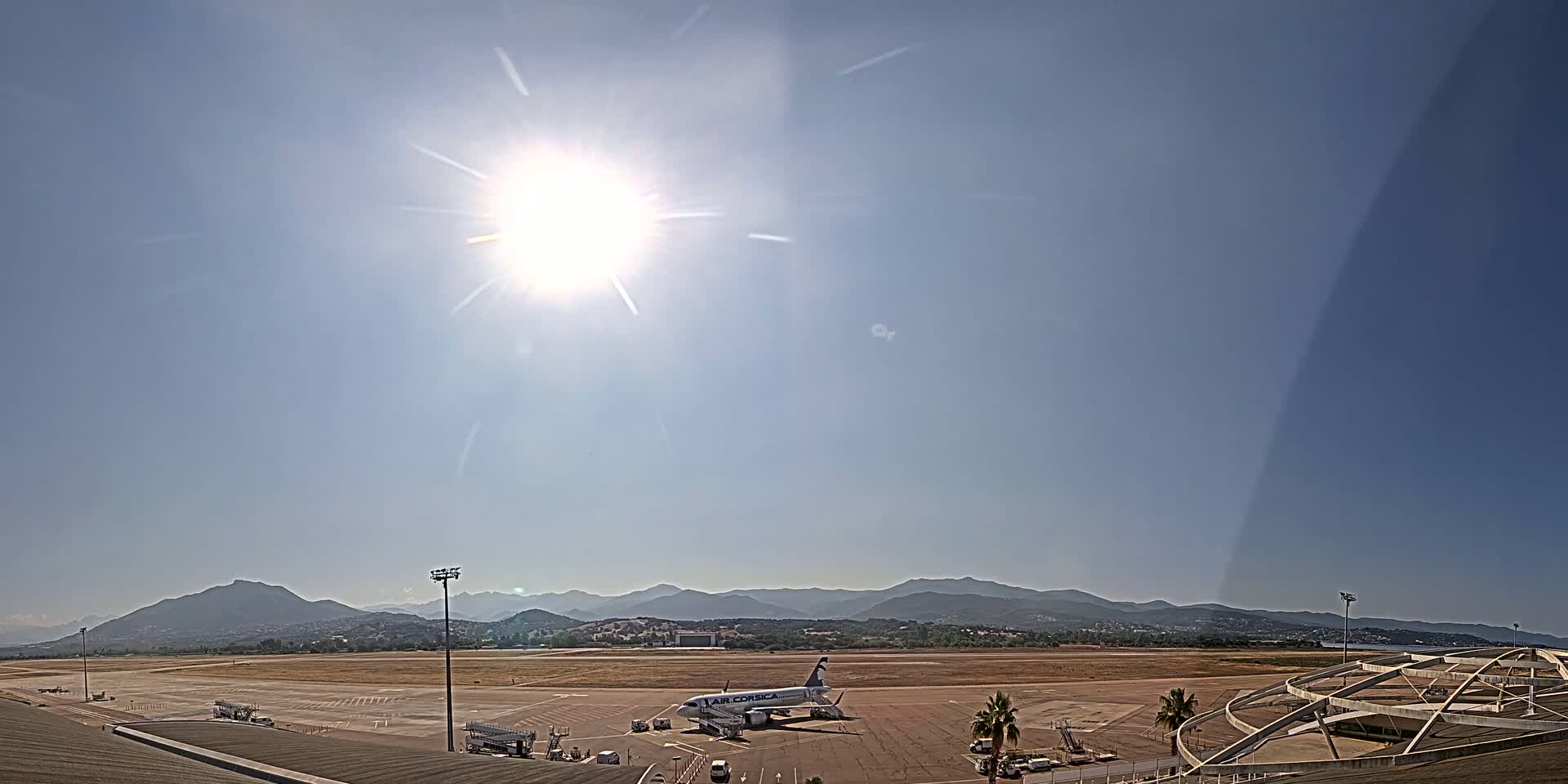 A passenger airplane sits on an airport tarmac under a bright, sunny sky, with mountains visible in the distance.