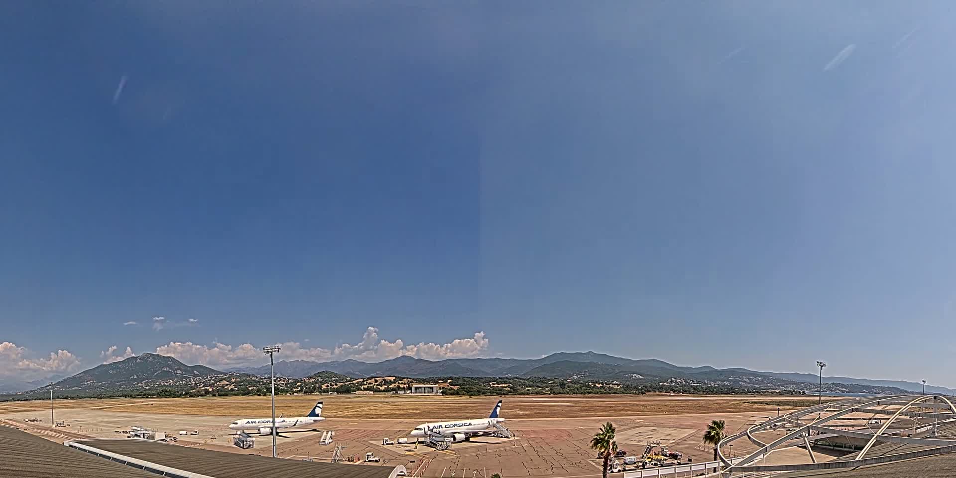 Two airplanes sit on a tarmac at an airport under a mostly clear blue sky, with mountains visible in the distance.