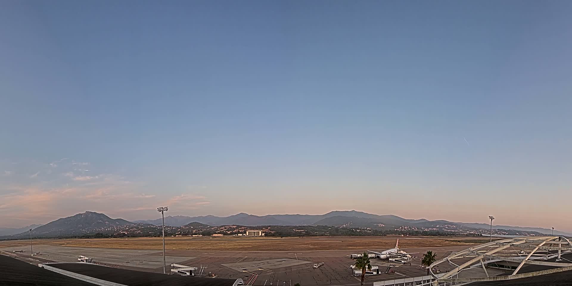 An airport tarmac with a plane and service vehicles is visible, under a clear, pale blue sky with a distant mountain range in the background.