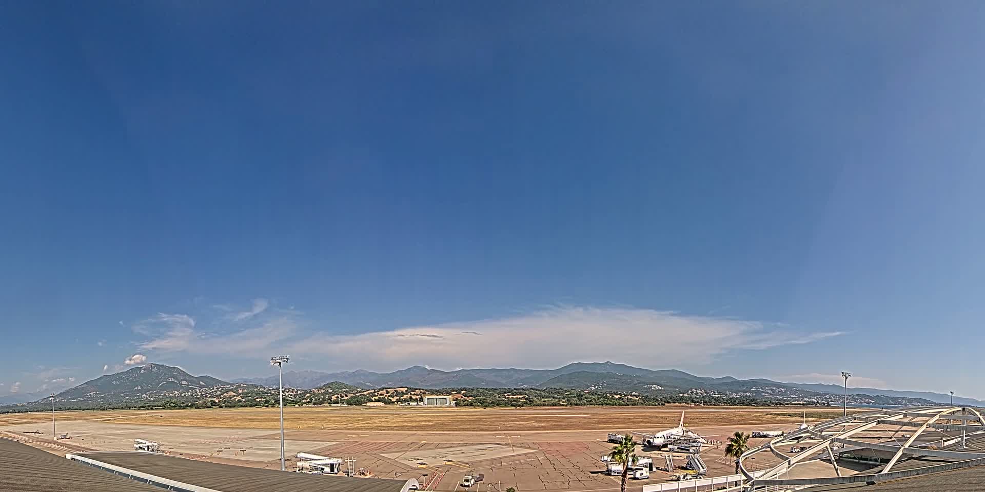 An airport tarmac with a few airplanes and support vehicles is visible under a mostly clear blue sky, with mountains in the background.