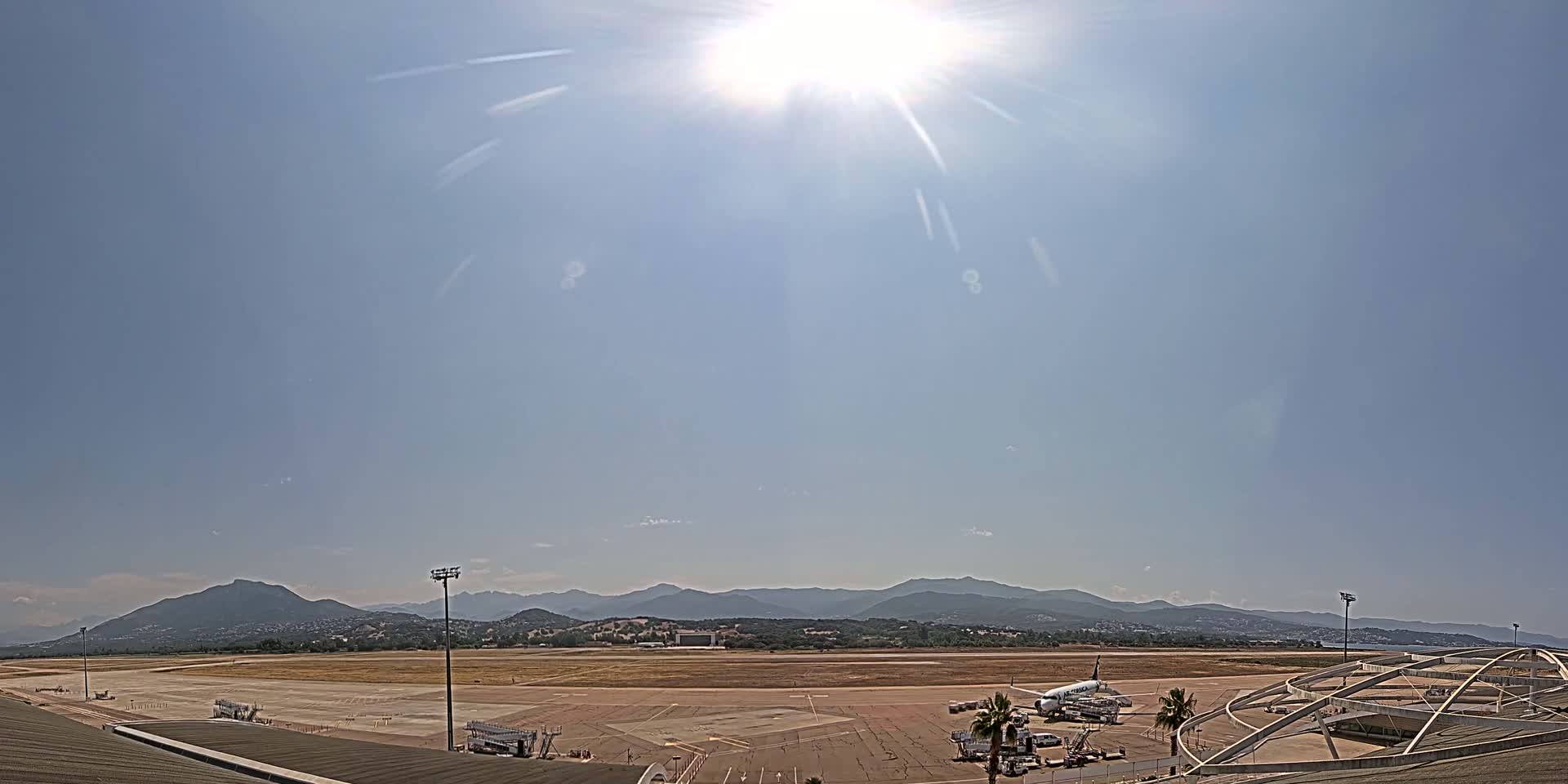 A sunny day reveals an airport tarmac with a plane parked near a terminal building, mountains visible in the distance.