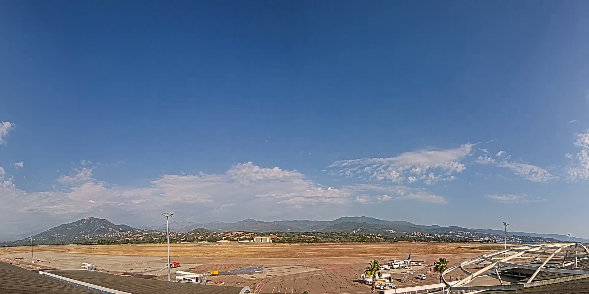An airport tarmac with several airplanes and support vehicles is visible under a mostly clear blue sky with scattered clouds, mountains in the background.