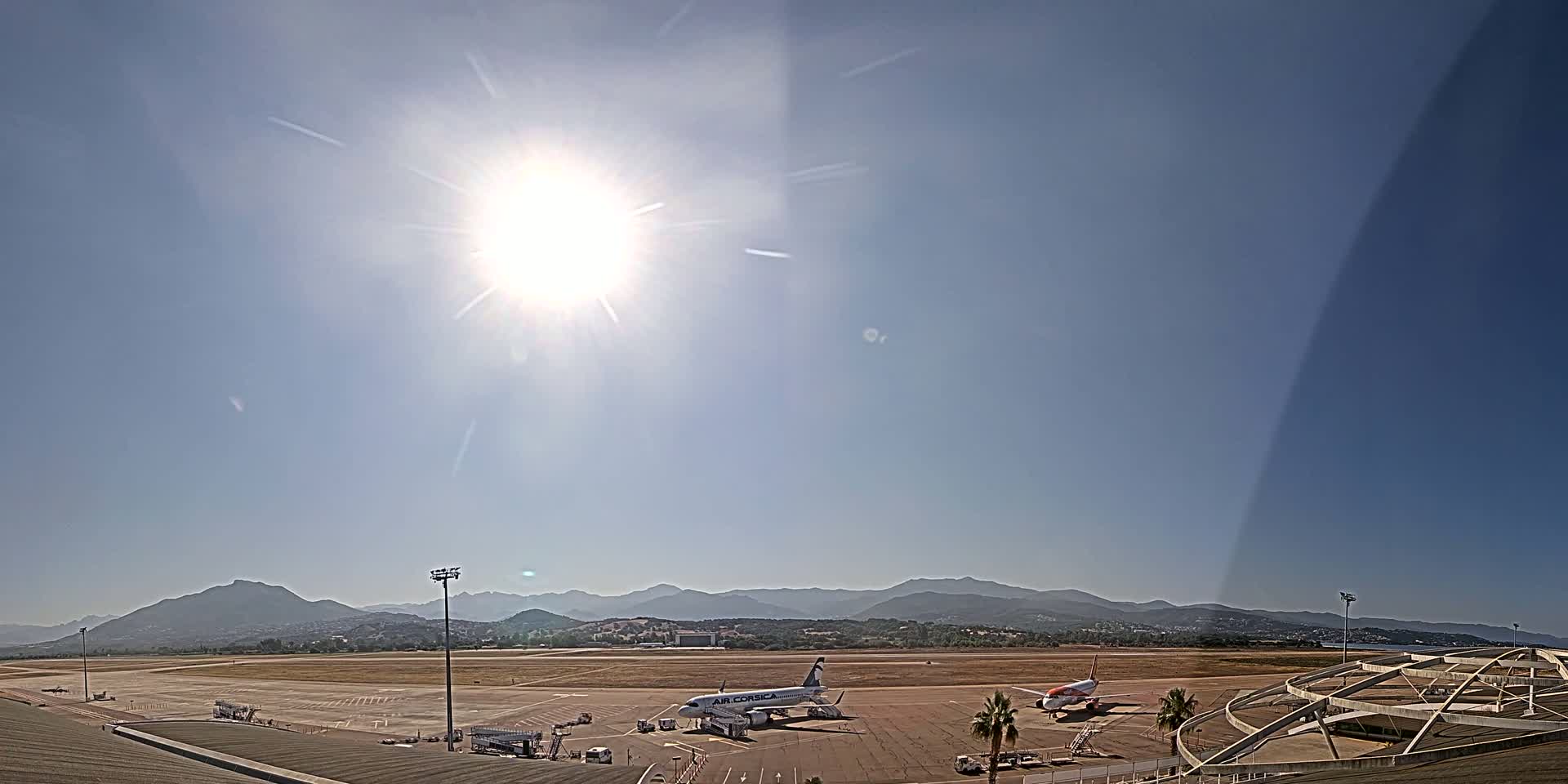 Two airplanes are parked on a sunny tarmac at an airport, with mountains visible in the distance under a clear sky.