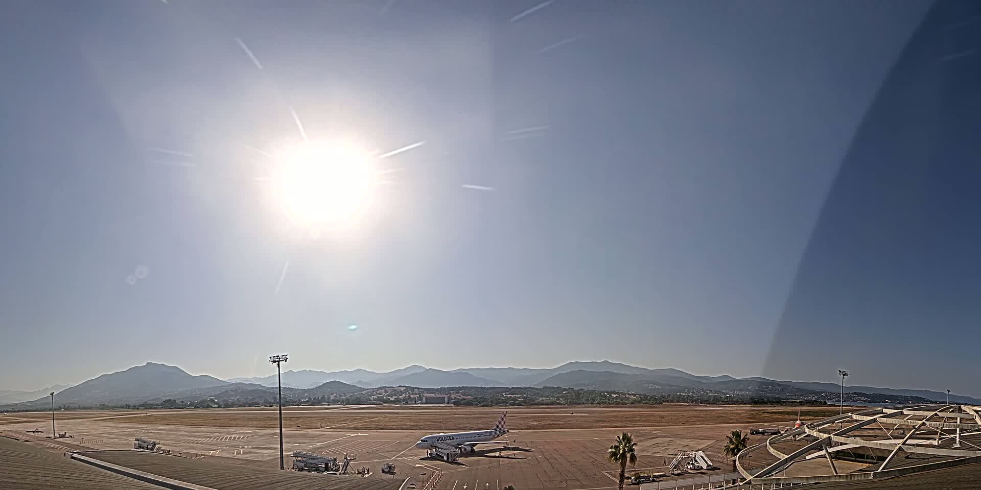 A passenger airplane sits on a tarmac at an airport on a sunny day with mountains in the background.