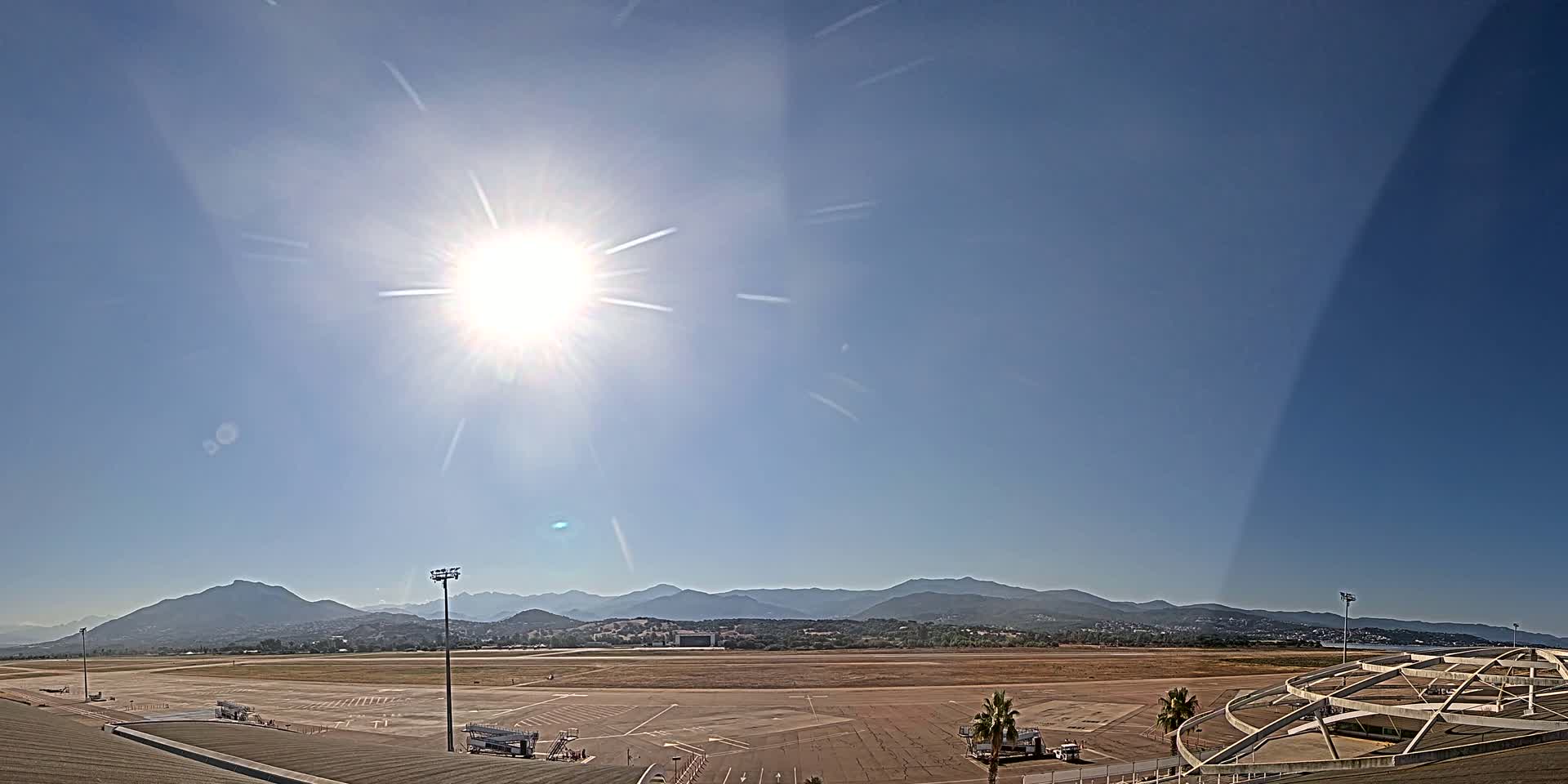 A sunny day reveals a wide expanse of airport tarmac, service vehicles, and distant mountains under a bright, clear sky.
